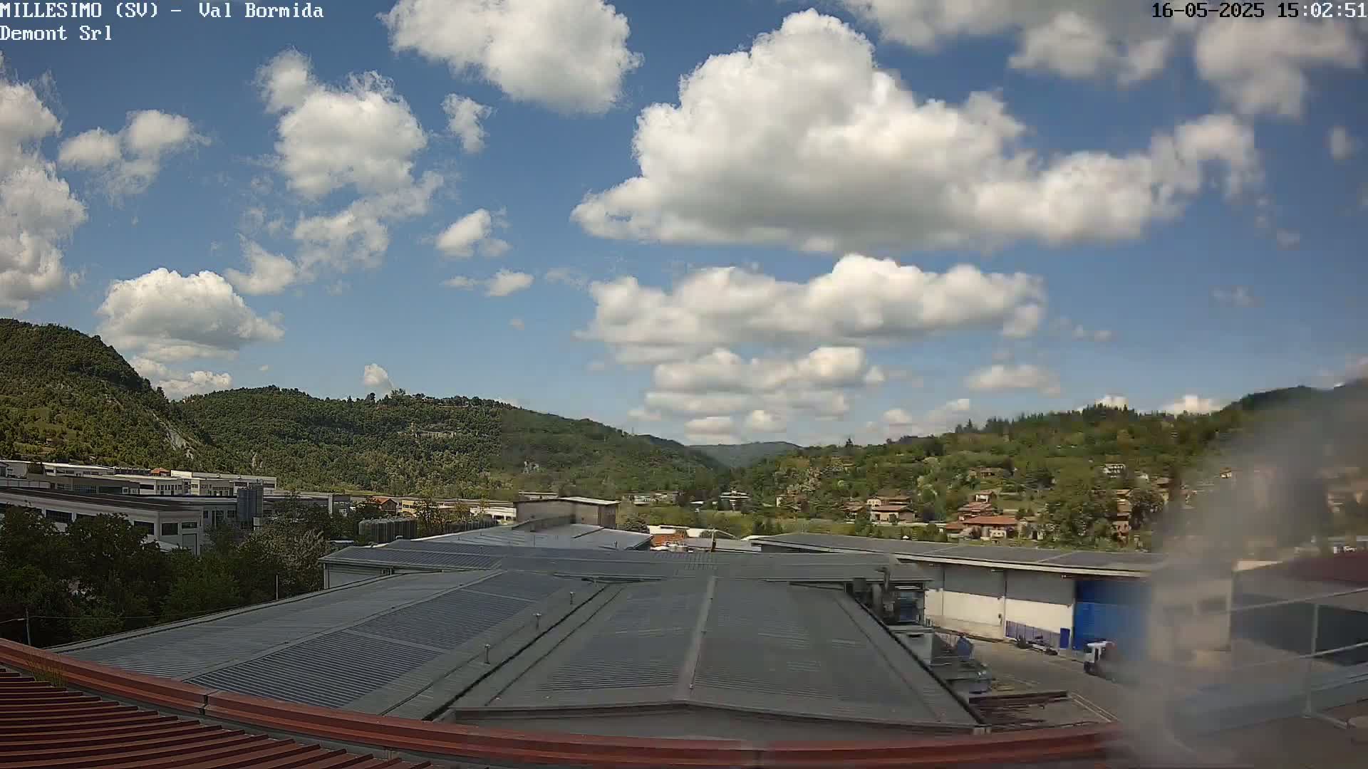 A partly cloudy day reveals a view of industrial buildings and houses nestled amongst green hills.
