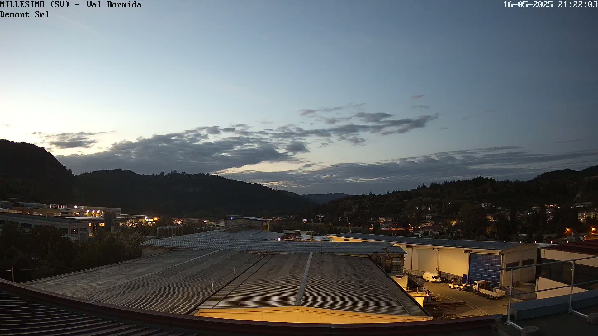 An industrial area with factory buildings is situated beneath a twilight sky with some clouds over a hilly landscape.