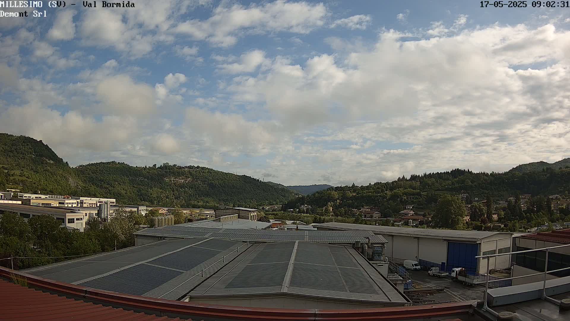 A partly cloudy sky overlooks a valley of green hills and industrial buildings, with several vehicles parked in a yard.