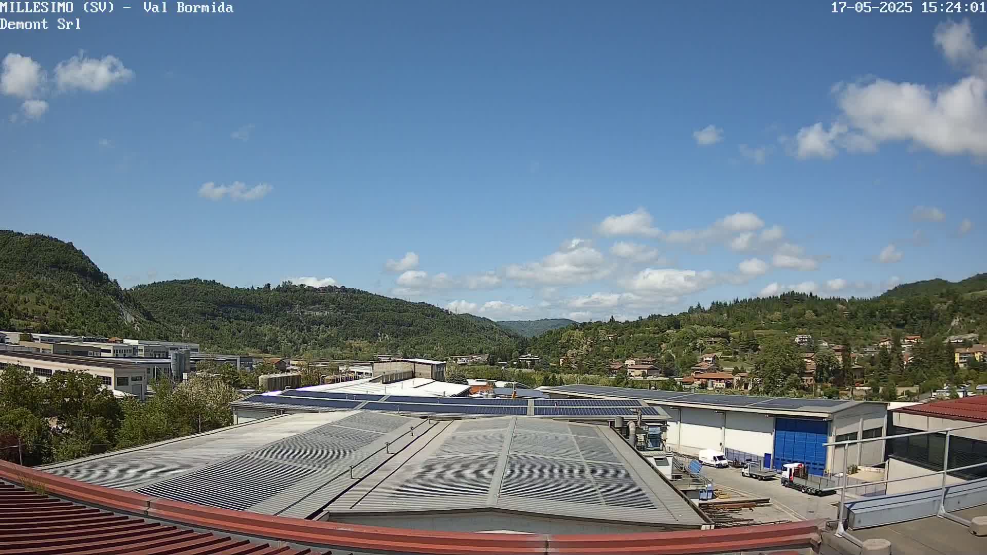 A view of a factory with solar panels on its roof, nestled in a valley surrounded by green hills under a mostly sunny sky with some scattered clouds.