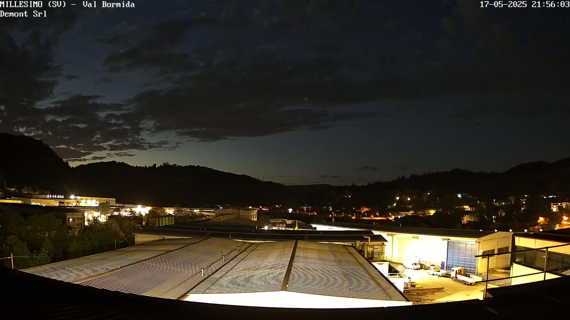 A nighttime view of an industrial area nestled in a valley under a partly cloudy sky.