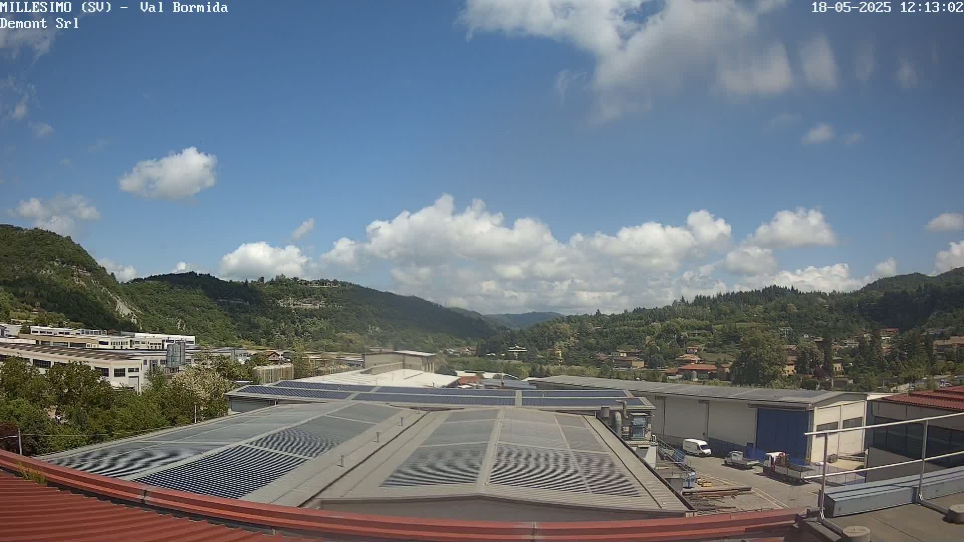 A partly cloudy sky overlooks industrial buildings and a hilly, green landscape.