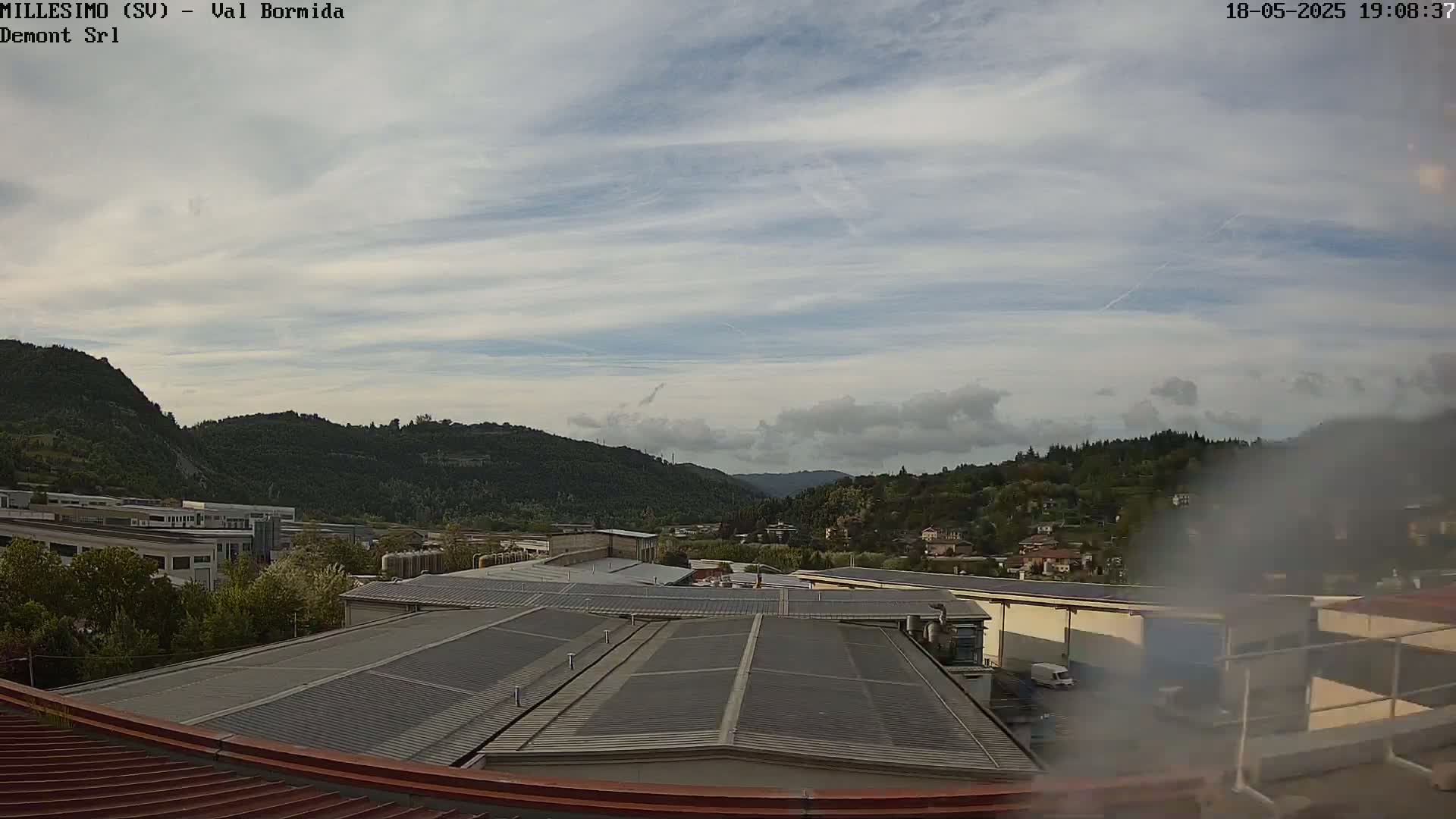 A view from a rooftop shows industrial buildings and a hazy, partly cloudy sky over rolling green hills.