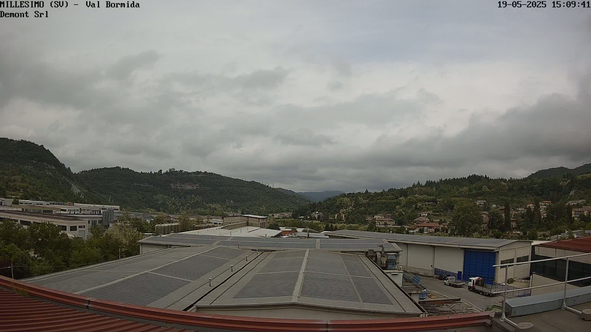 A view from a rooftop shows overcast skies over a valley with industrial buildings and residential houses nestled among green hills.