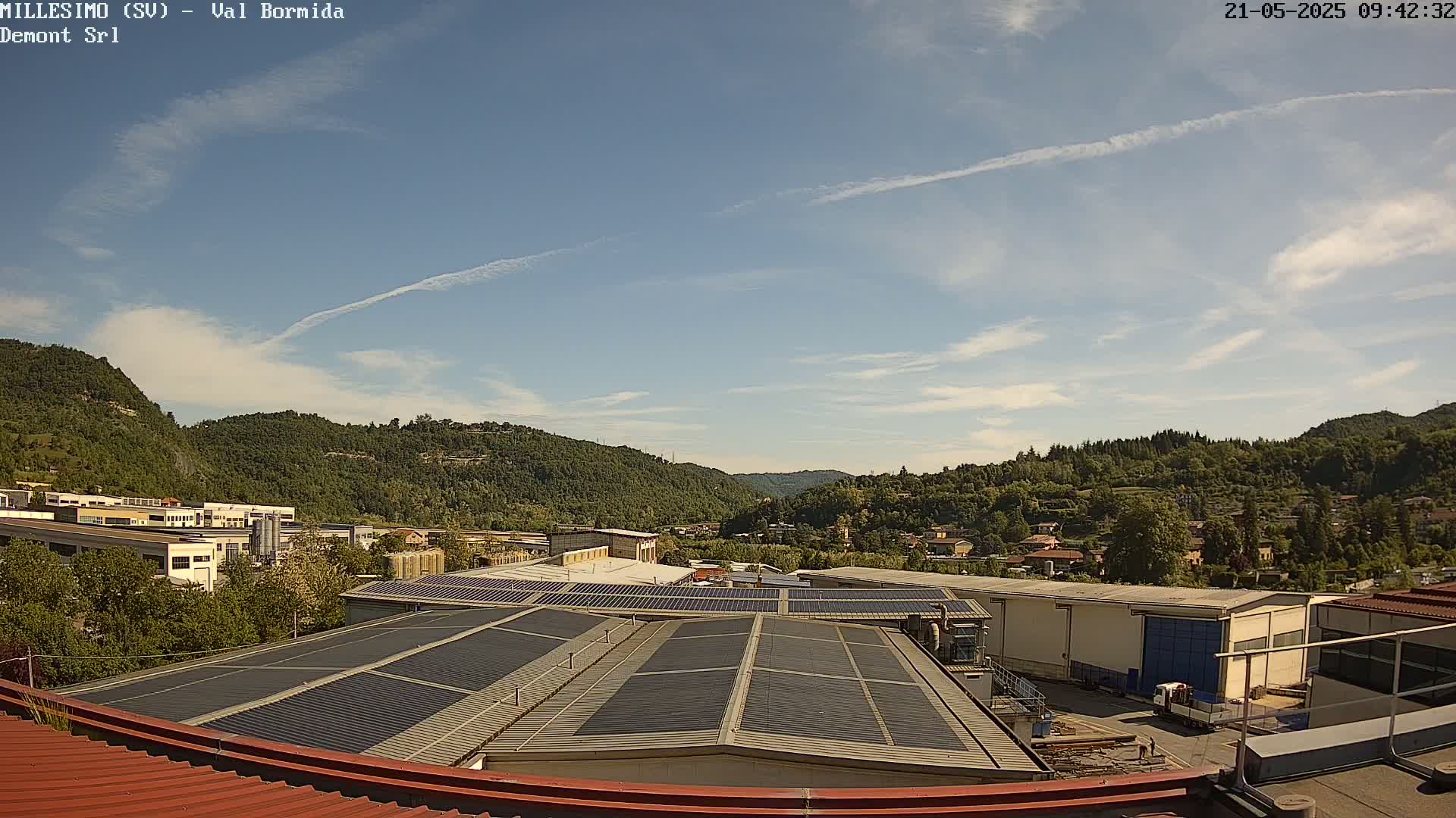 A mostly sunny day overlooks a valley of green hills and industrial buildings with solar panels on the roofs.