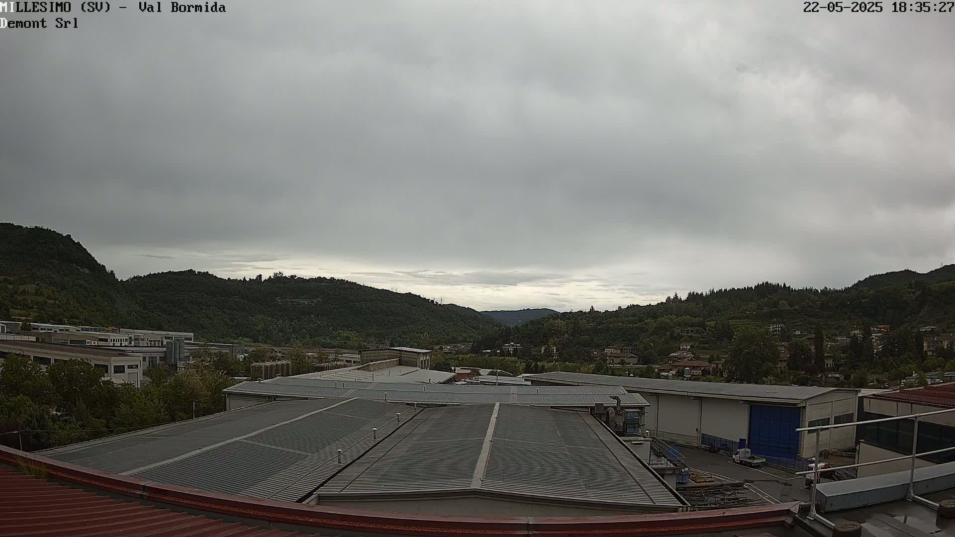A view from a rooftop shows overcast skies over a valley filled with green hills and industrial buildings.