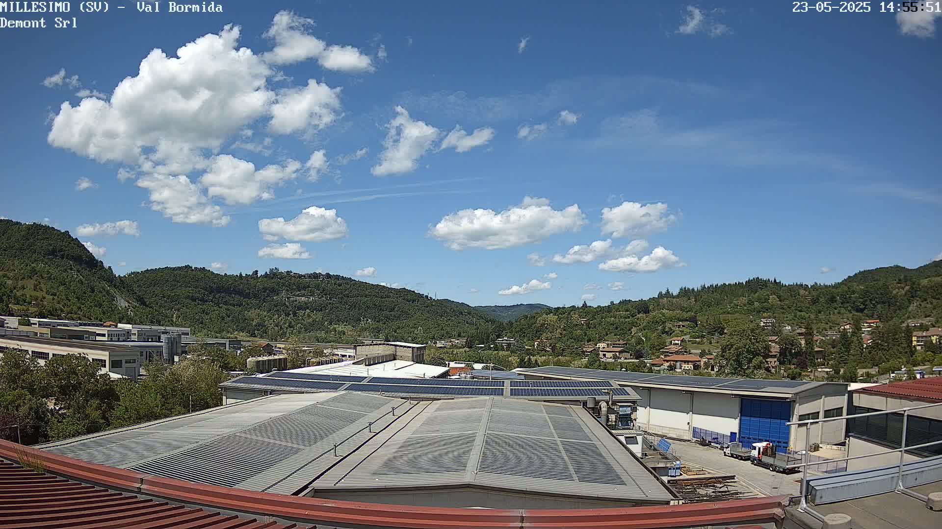 A partly cloudy blue sky overlooks an industrial area nestled in a valley between green hills.