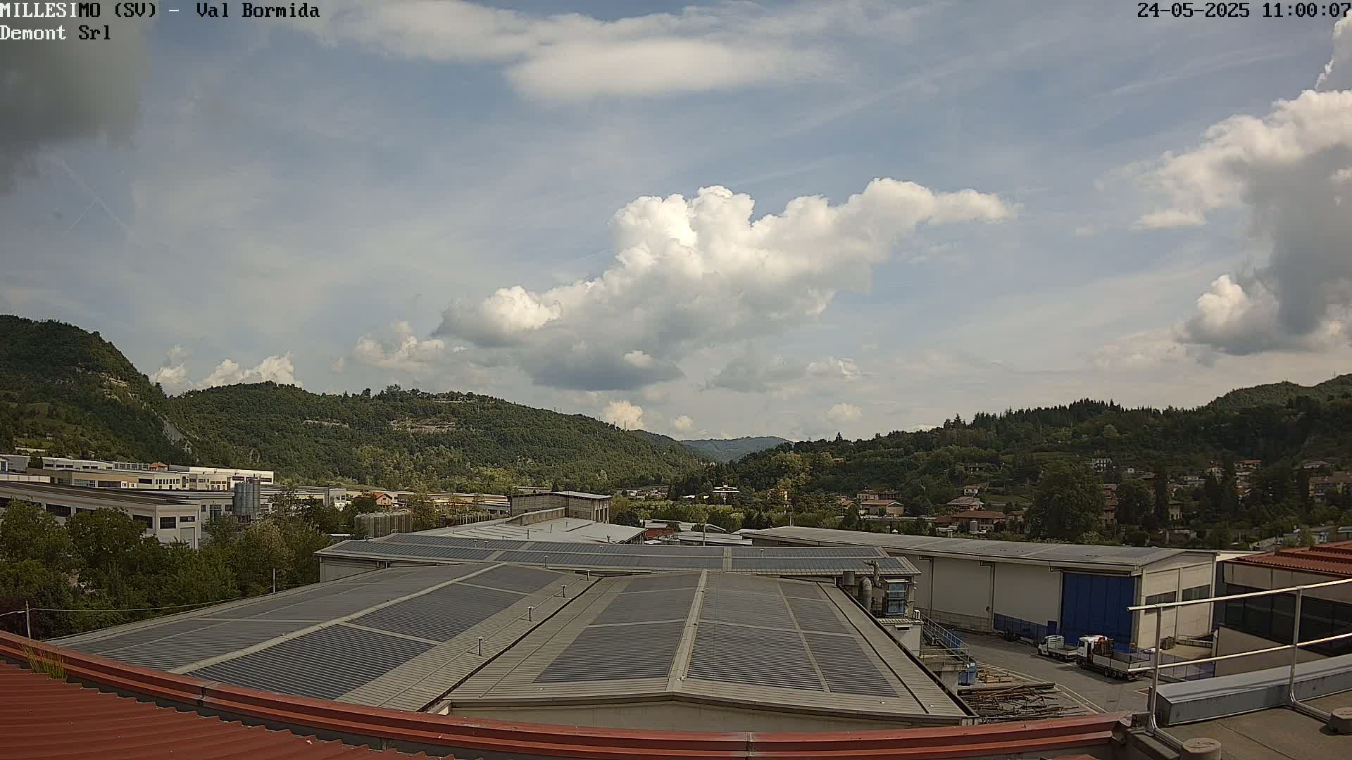 A partly cloudy sky overlooks a valley of green hills and industrial buildings, viewed from a rooftop with solar panels.