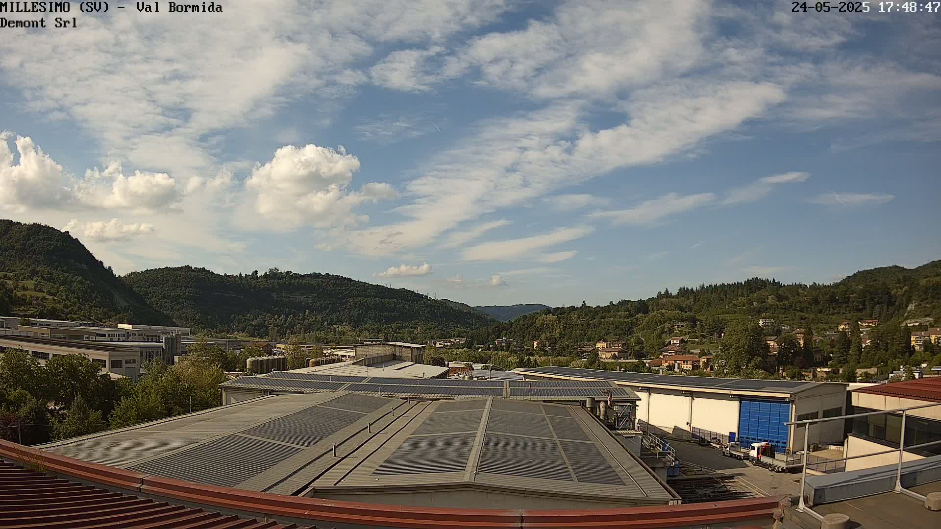 A partly cloudy sky overlooks a factory complex with solar panels on its roofs, nestled in a valley surrounded by green hills.