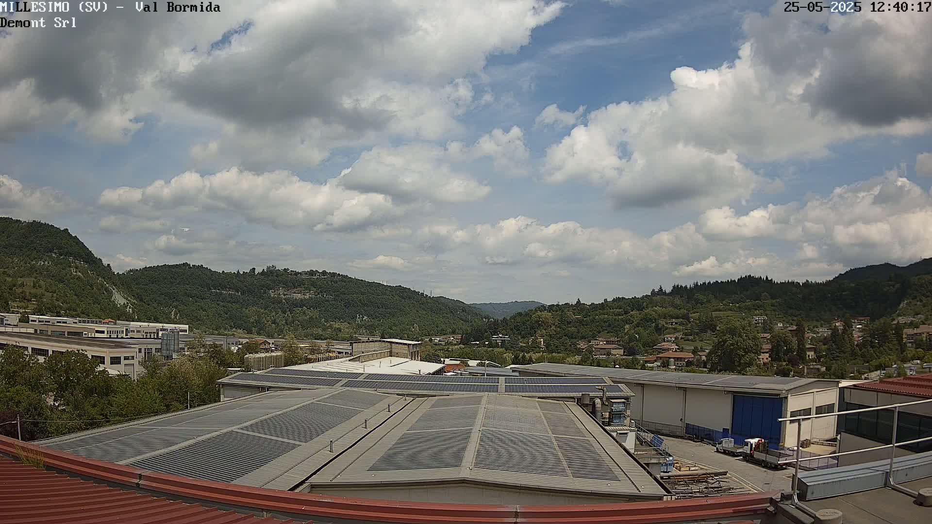 A partly cloudy sky overlooks a valley of green hills and industrial buildings with solar panels on their roofs.