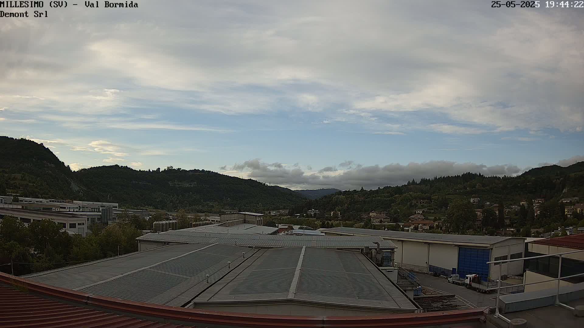 A view from a rooftop shows industrial buildings and a residential area nestled in a valley under a partly cloudy sky.