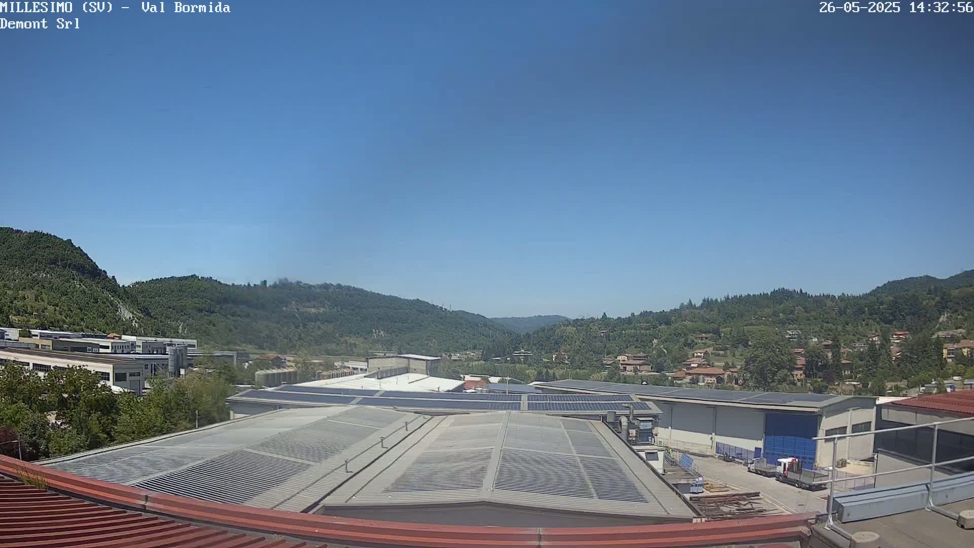 A clear blue sky overlooks an industrial area with solar panels on rooftops, nestled in a valley surrounded by green hills.