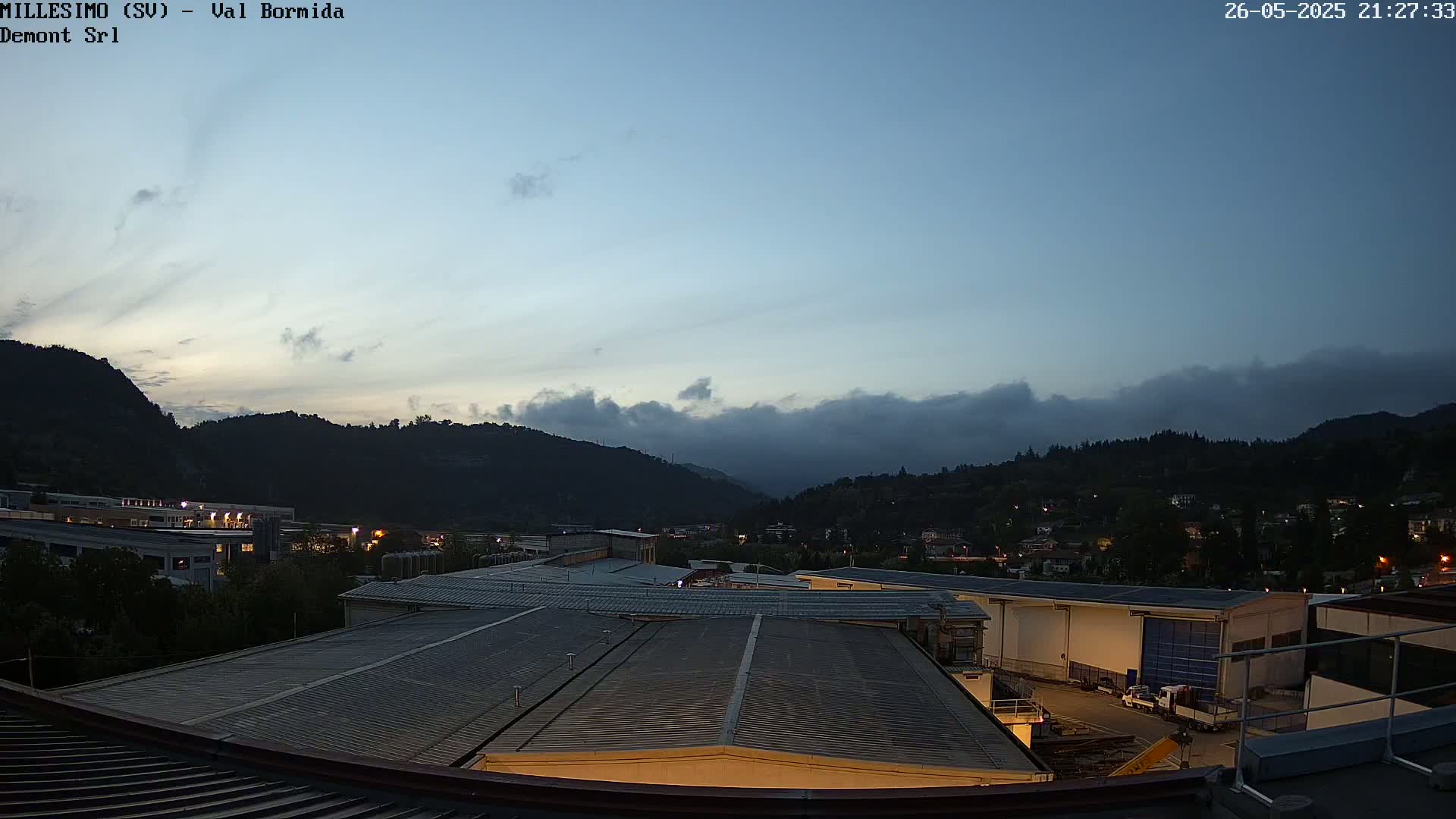 A view from a rooftop shows a twilight sky with some clouds over a valley of buildings and hills.
