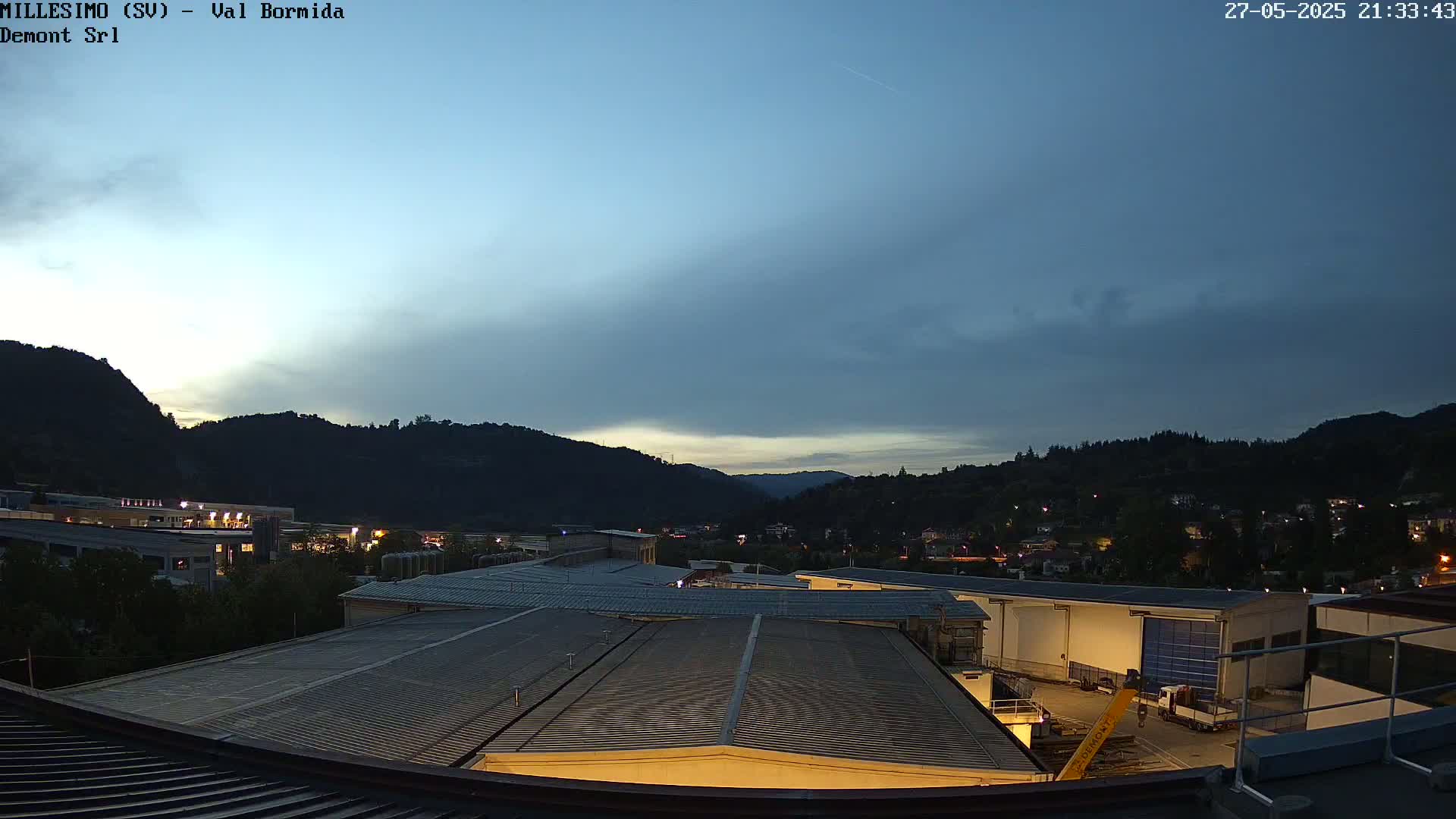 An industrial area nestled in a valley between hills is visible at twilight under a mostly clear sky.