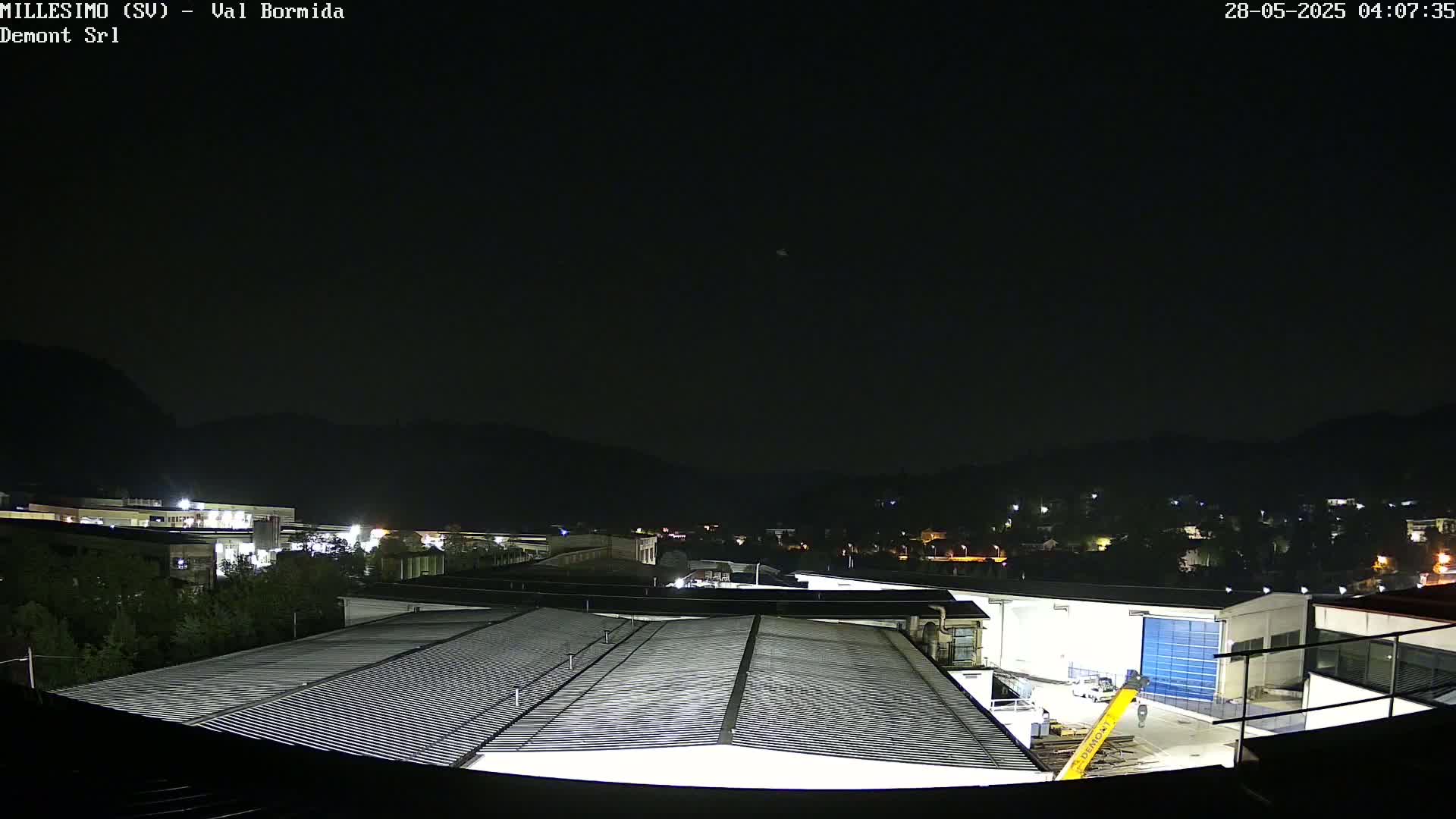 A nighttime view of industrial buildings and a small town nestled in dark hills under a clear, dark sky.