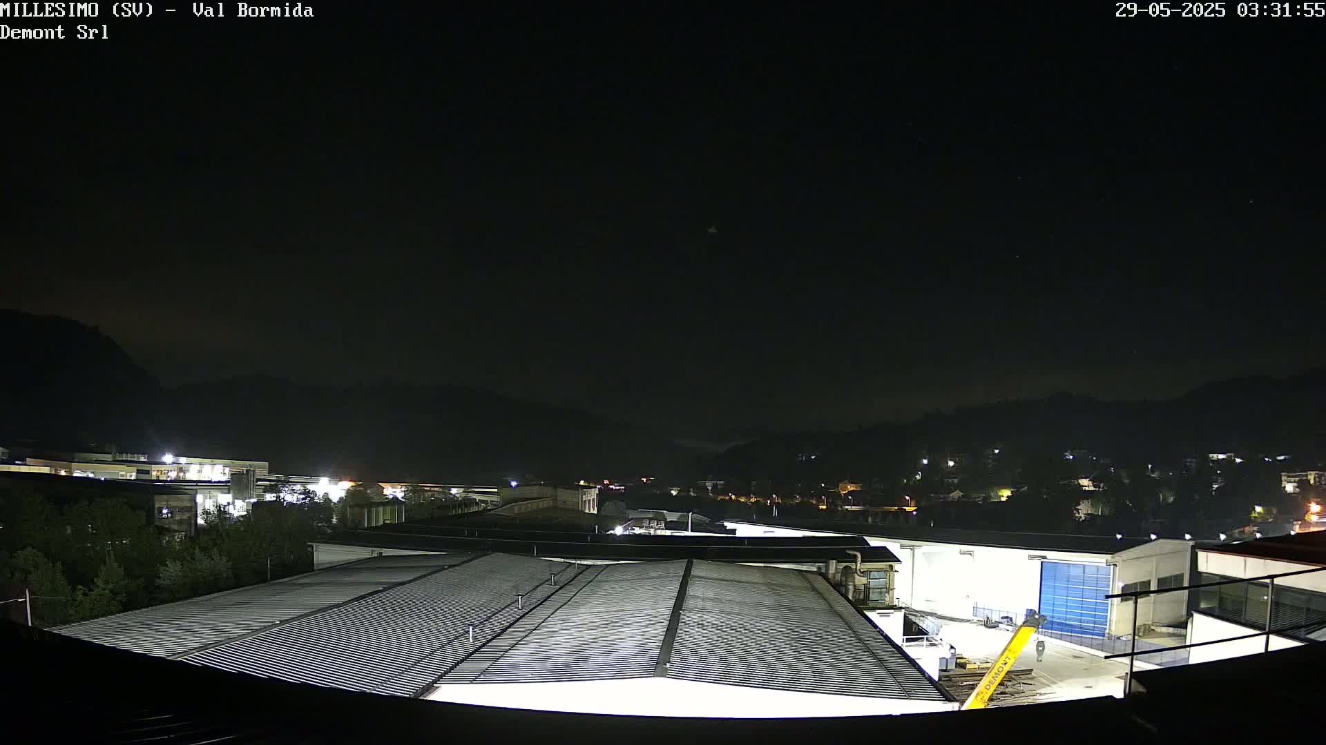 A nighttime view of an industrial area with a corrugated metal roof in the foreground, under a clear, dark sky, shows some distant lights on a hillside.