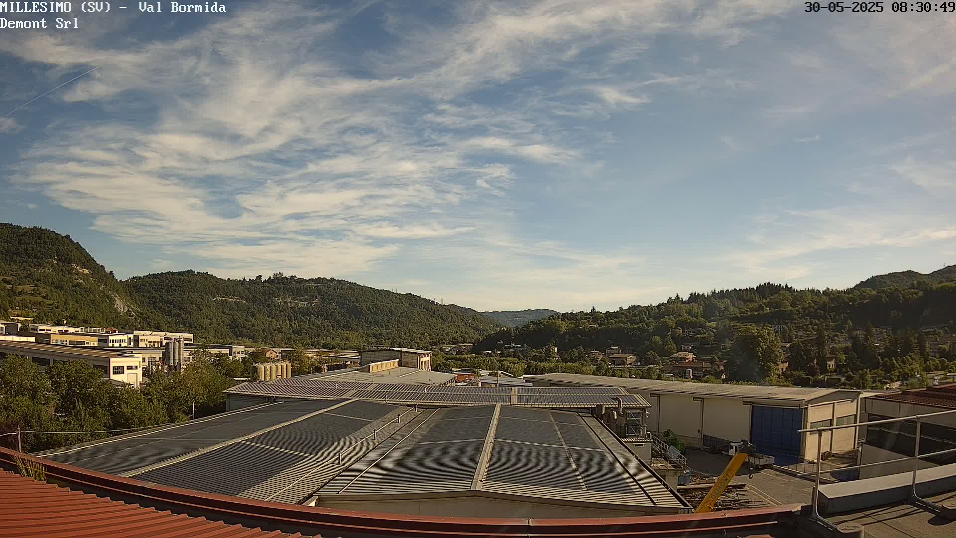 A mostly sunny day reveals a view of industrial buildings with solar panels on their roofs, nestled in a valley surrounded by green hills.