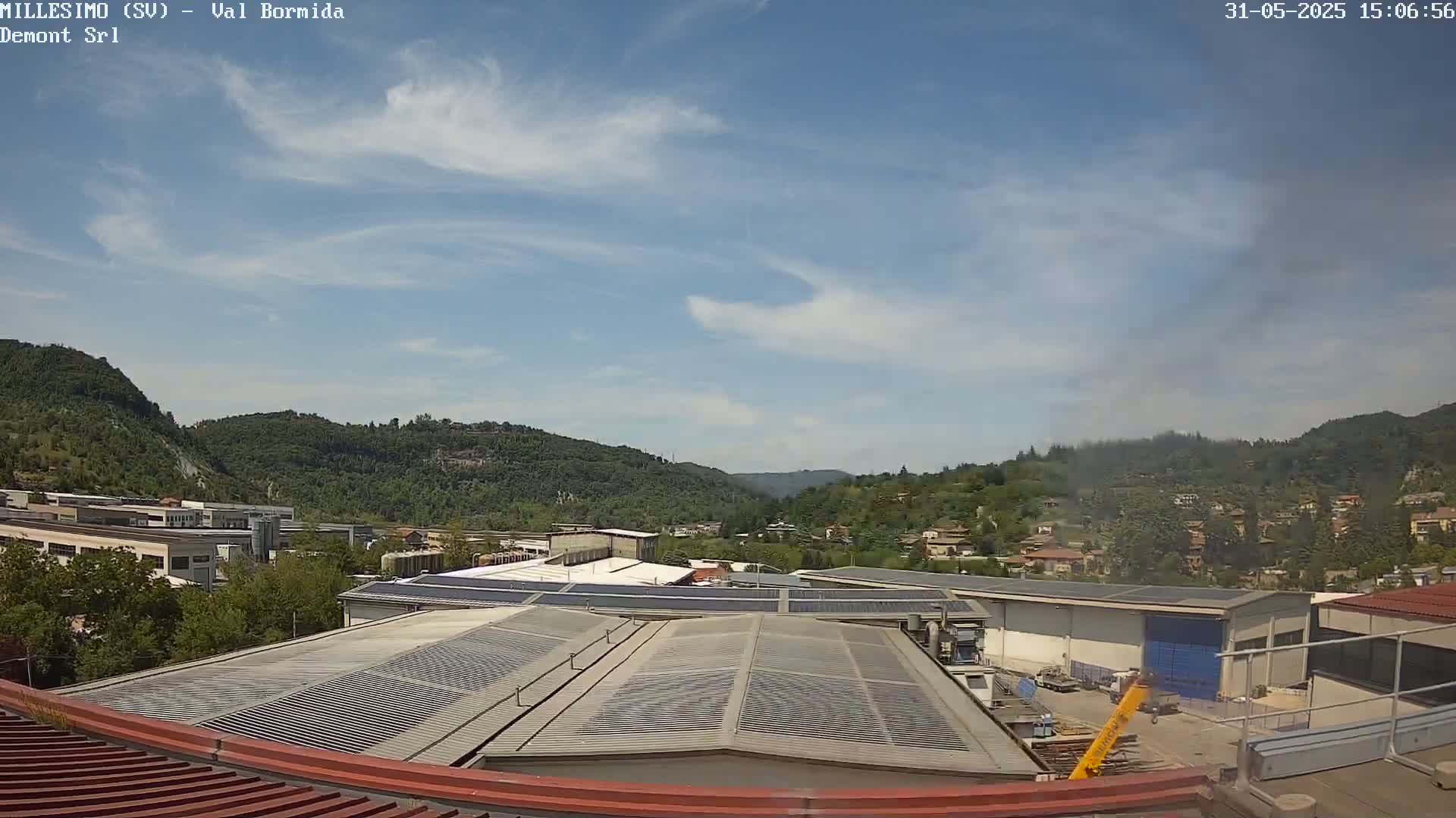 A mostly sunny day reveals a view of industrial buildings and a valley filled with green hills, under a partly cloudy sky.