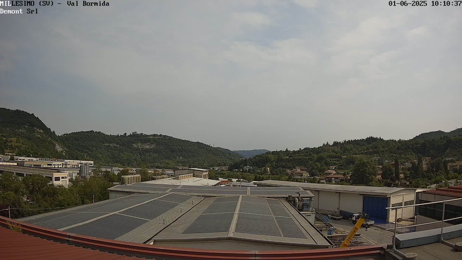 A mostly cloudy day reveals a view of industrial buildings with solar panels on their roofs, set against a backdrop of rolling green hills and a small town.