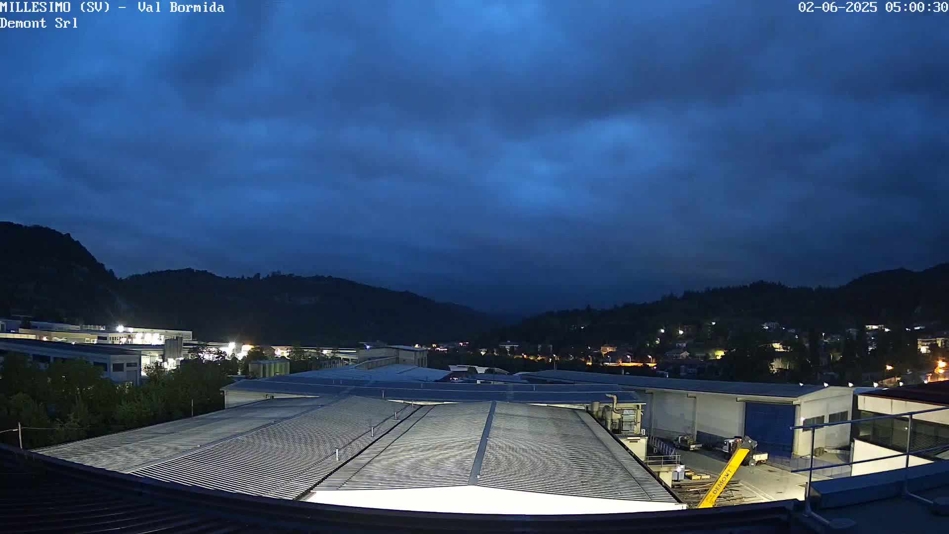 A view from a rooftop shows a factory complex under a dark blue, cloudy sky at night, with hills in the background.
