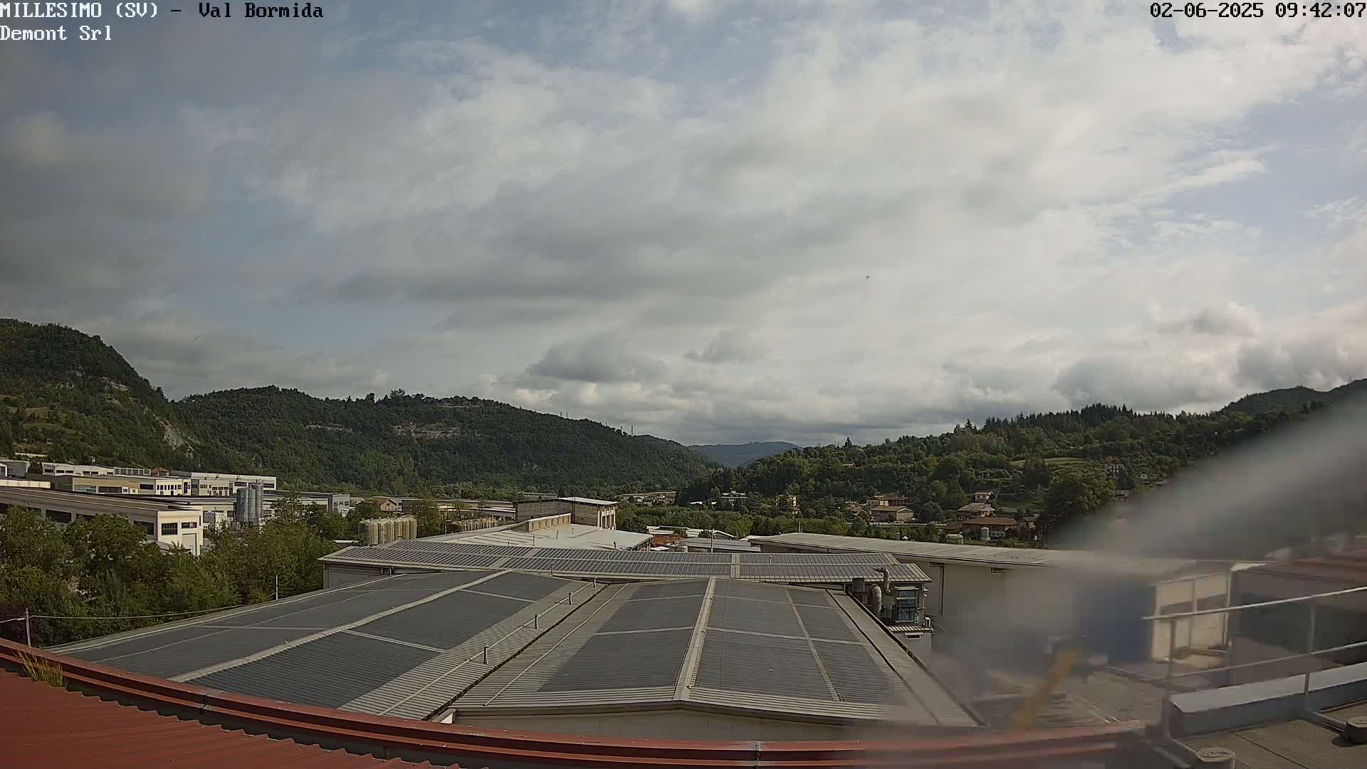 A view from a rooftop shows a partly cloudy sky over a valley of green hills and industrial buildings.