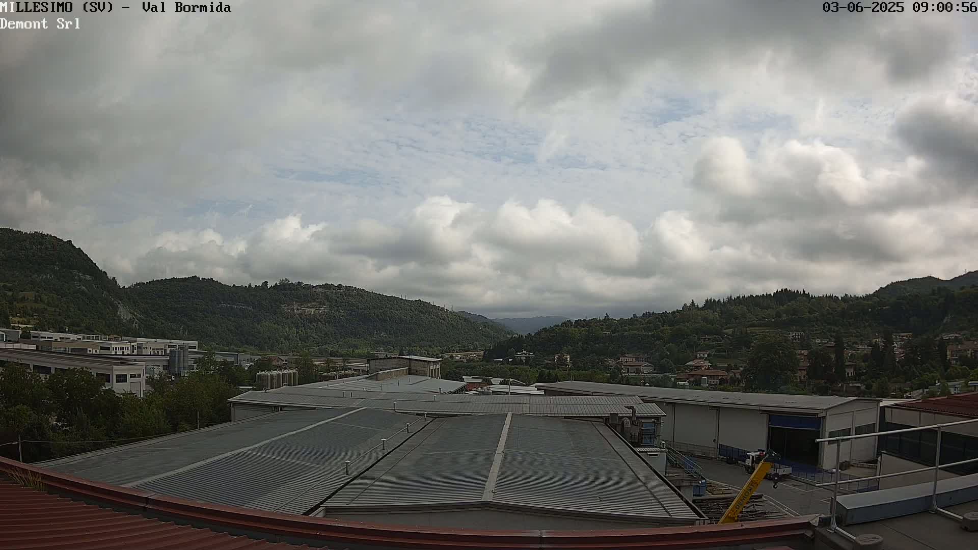 A mostly cloudy sky over a valley of green hills and industrial buildings, viewed from a rooftop.