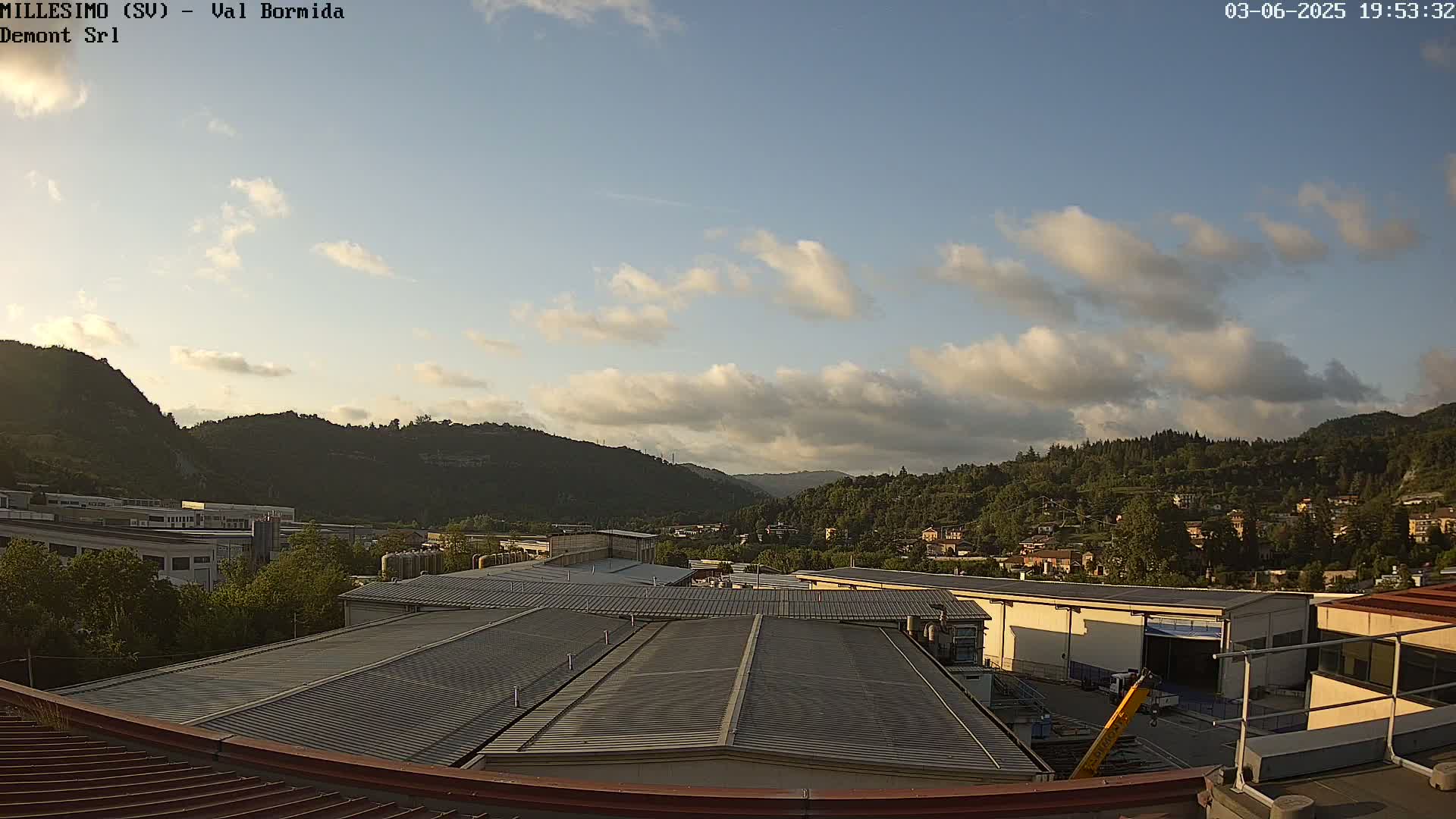 A view from a rooftop shows industrial buildings and a hilly landscape under a partly cloudy sky.