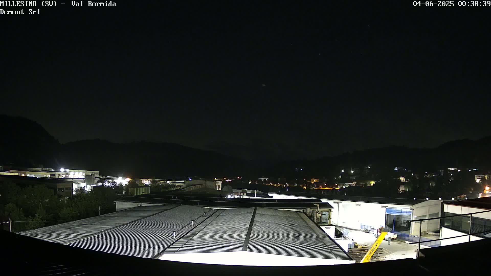A nighttime view of an industrial area with several buildings, hills in the background, and a clear, dark sky.