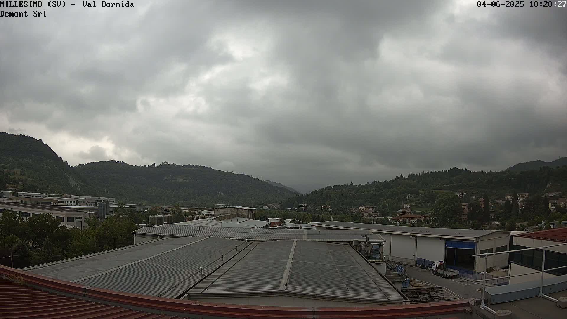Overcast skies loom over a valley of green hills and industrial buildings, with the foreground showing the corrugated metal roof of a factory.