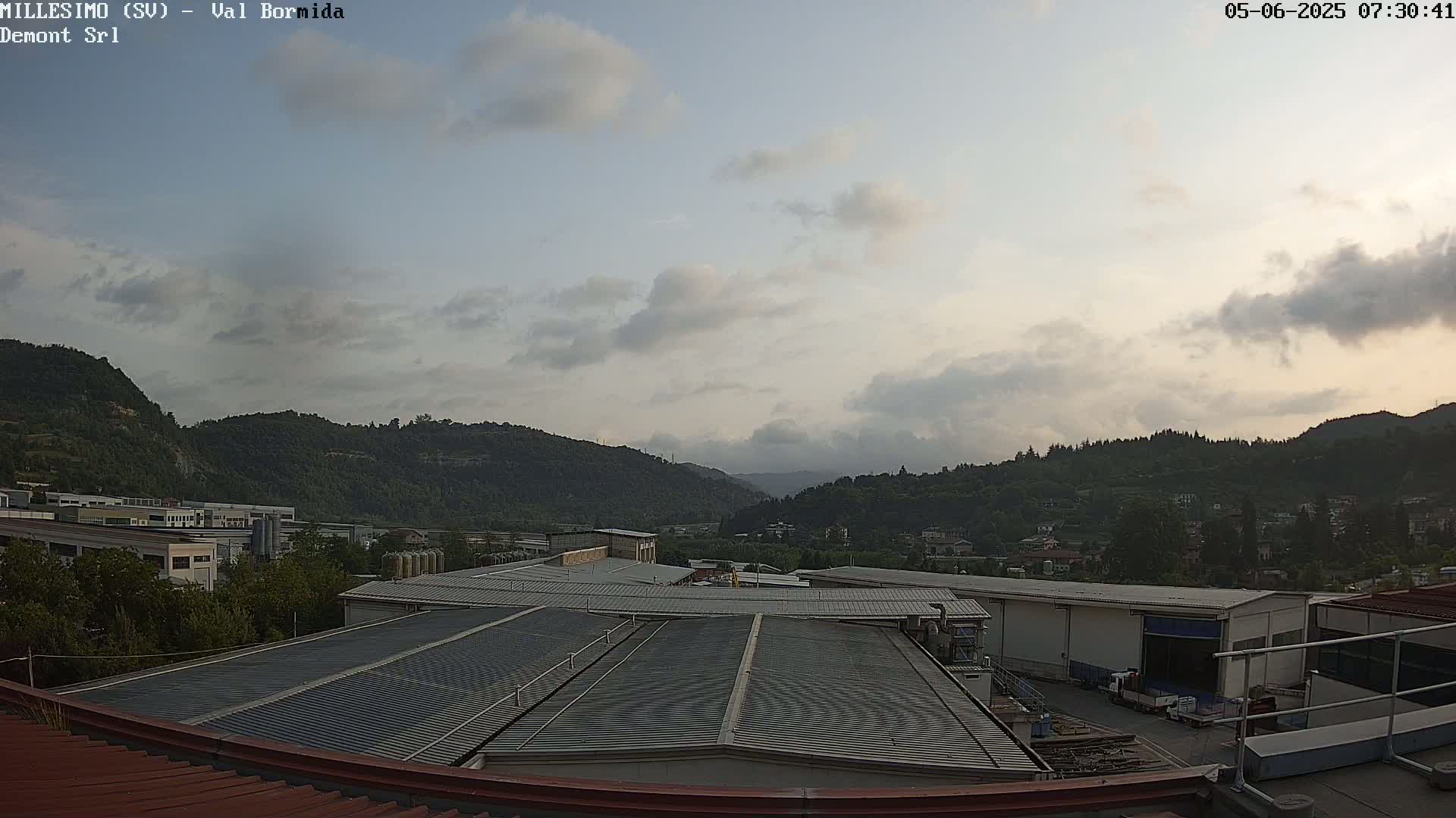 A view from a rooftop shows a partly cloudy sky over a valley filled with industrial buildings and hills covered in green vegetation.