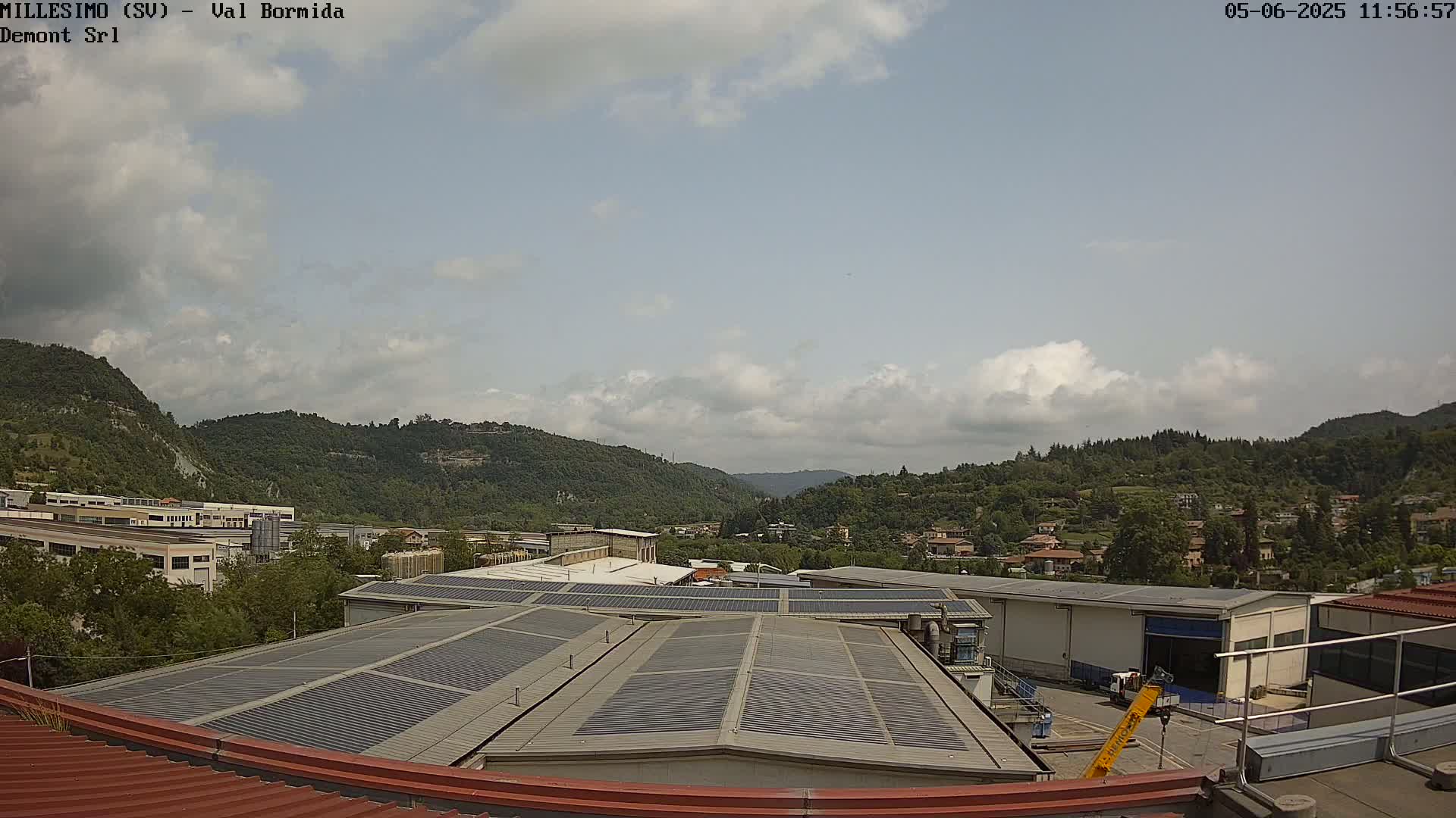 A partly cloudy sky overlooks a valley of green hills and industrial buildings with solar panels on their roofs.
