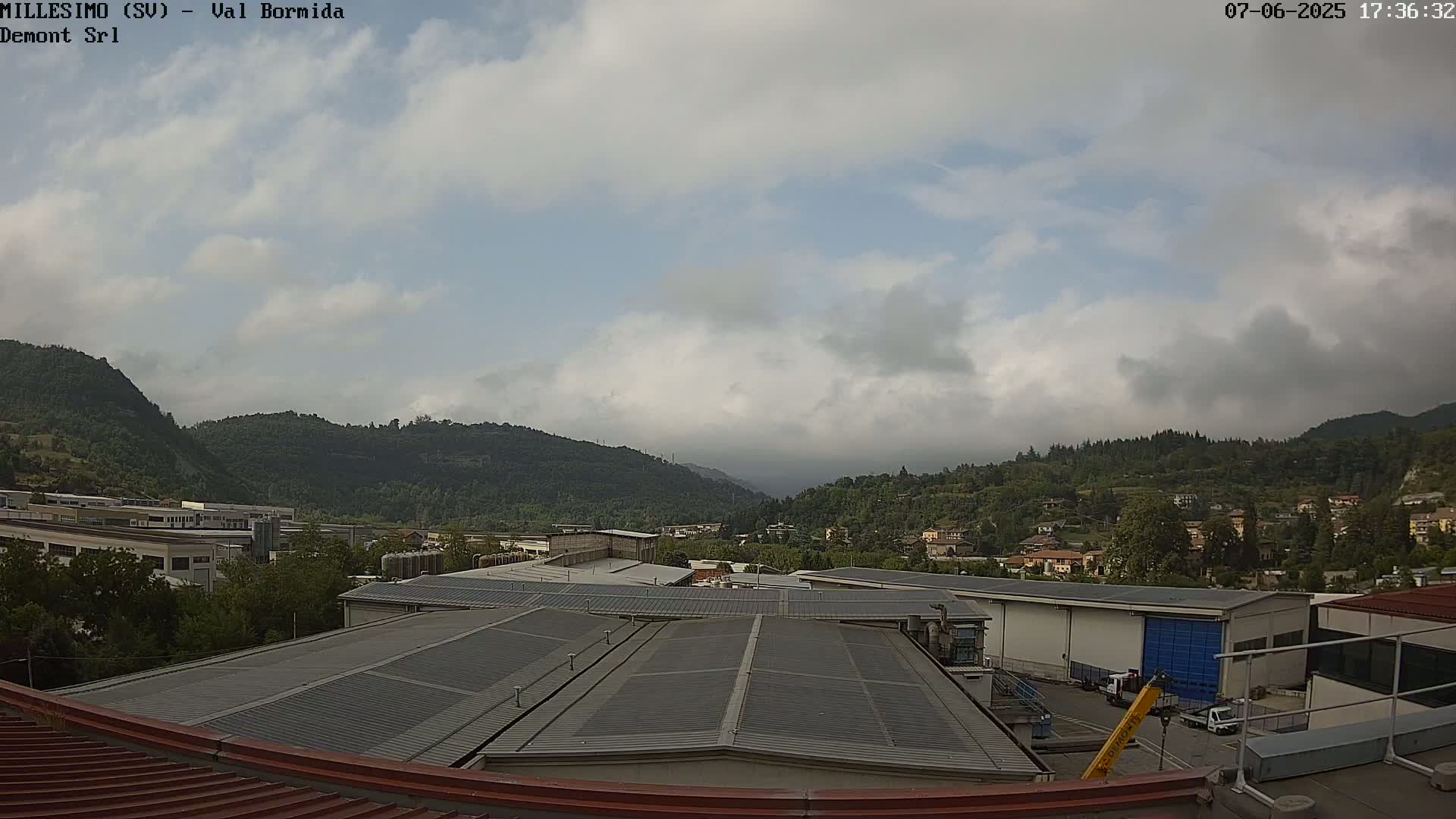 A cloudy sky overlooks a valley of green hills, industrial buildings, and a town nestled amongst them.