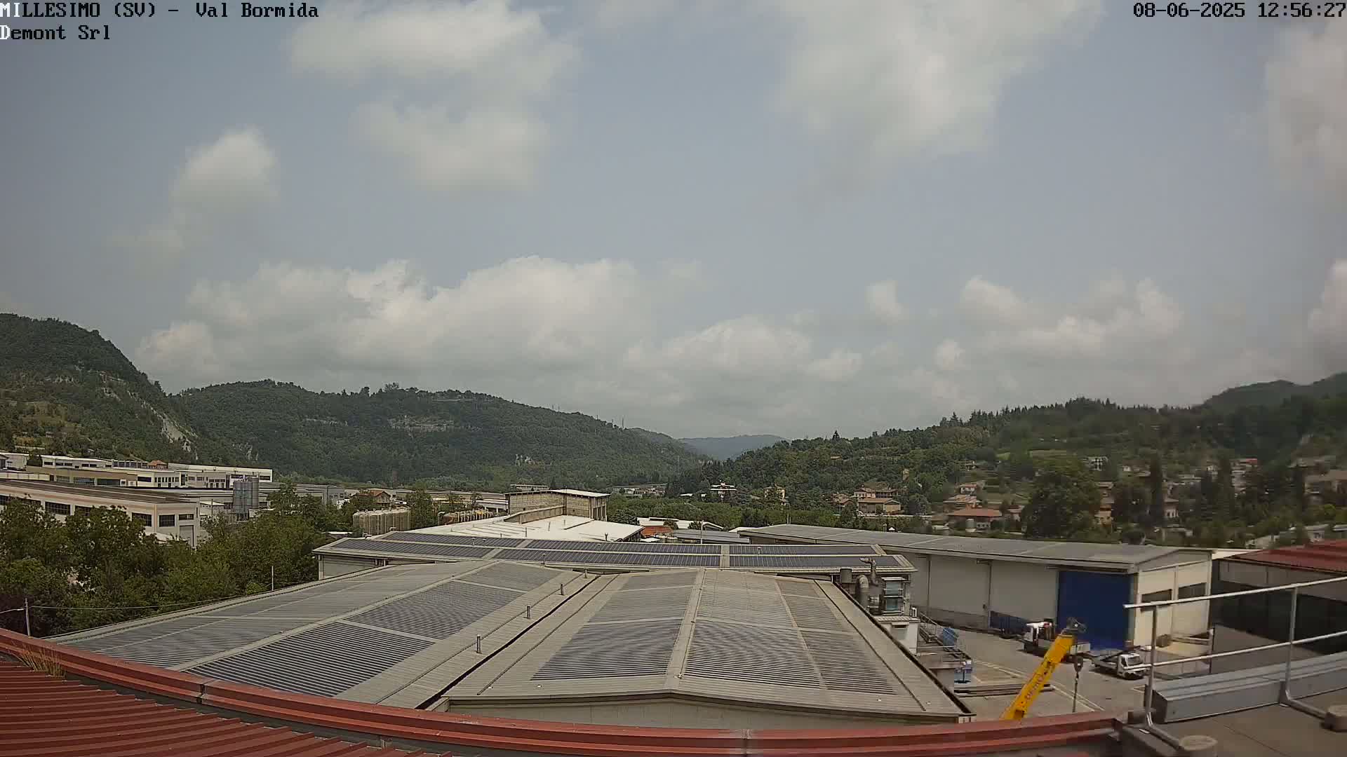 A partly cloudy sky overlooks industrial buildings and a valley of green hills and houses.