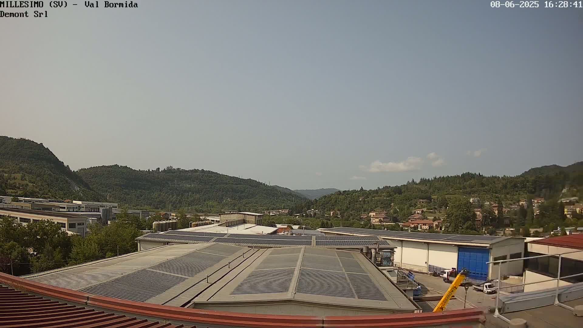 A view from a rooftop shows a mostly sunny day with light clouds over green hills and a valley containing industrial buildings and residential homes.