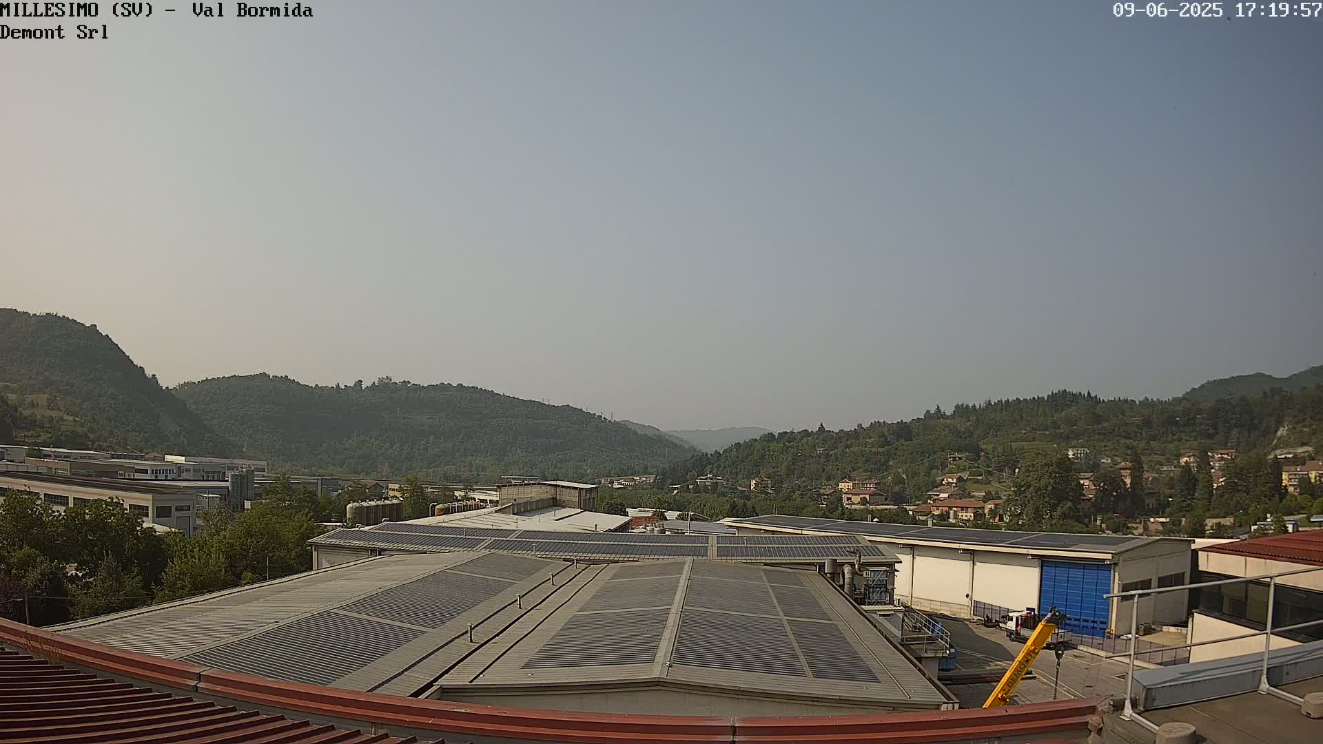 A hazy, sunny day overlooks a valley of green hills and industrial buildings with solar panels on their roofs.