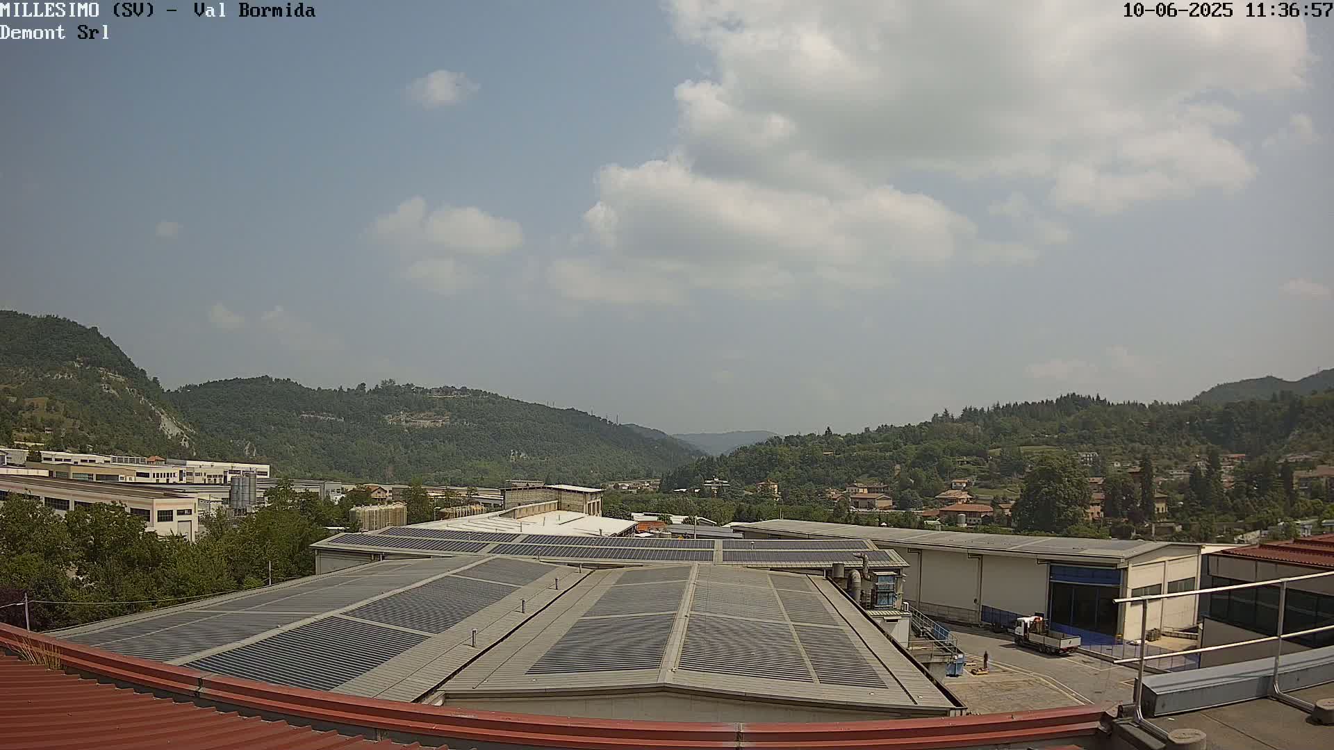 A partly cloudy sky overlooks an industrial area nestled in a valley between green hills.