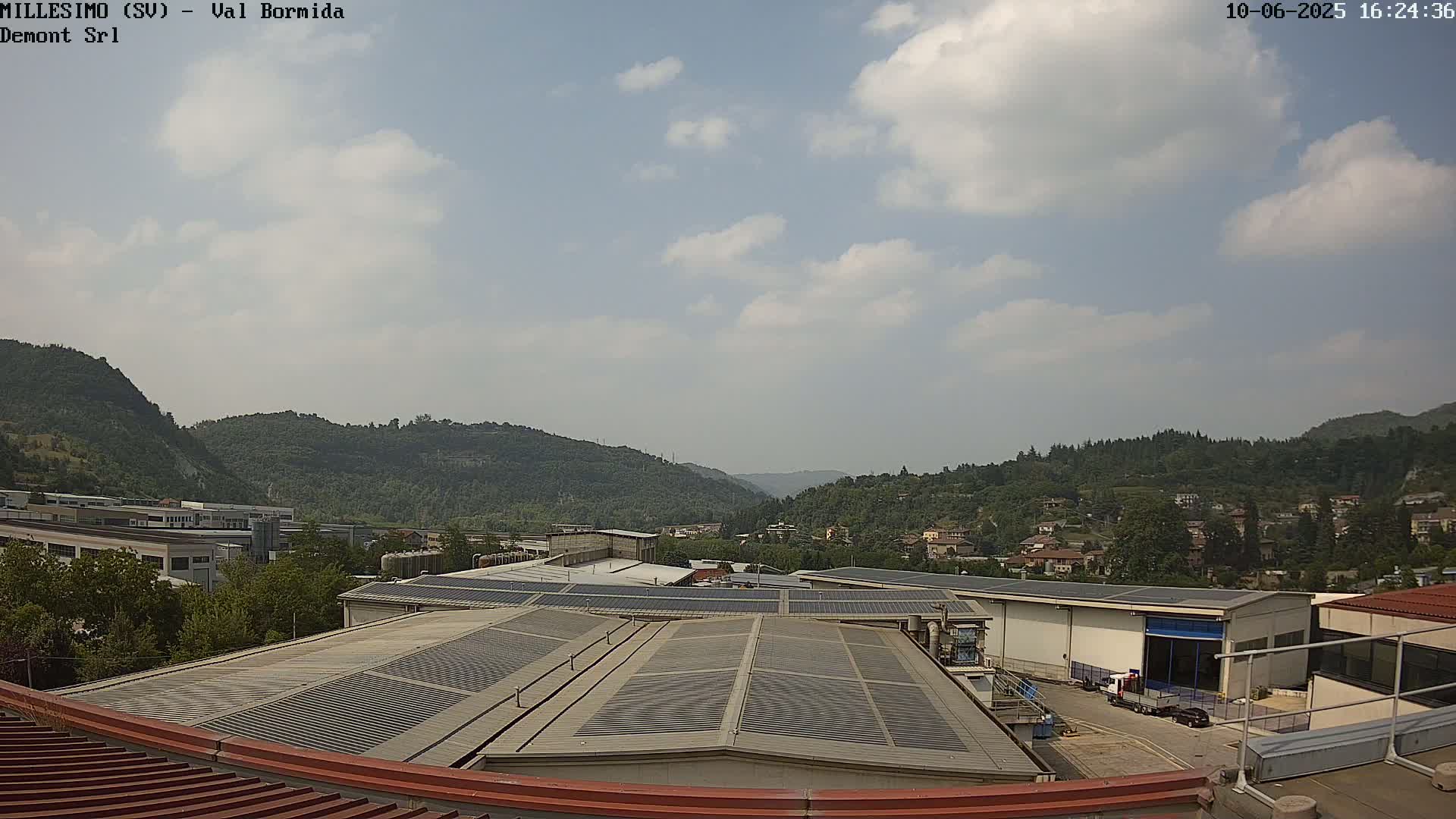 A partly cloudy day reveals a valley with industrial buildings and residential areas nestled amongst green hills.