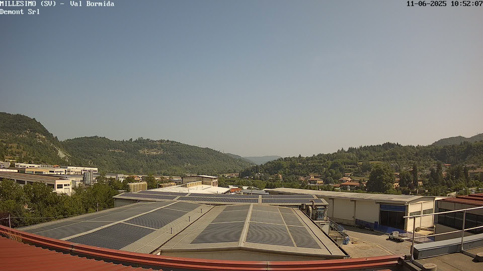A rooftop covered in solar panels overlooks a valley of green hills and industrial buildings under a clear, sunny sky.