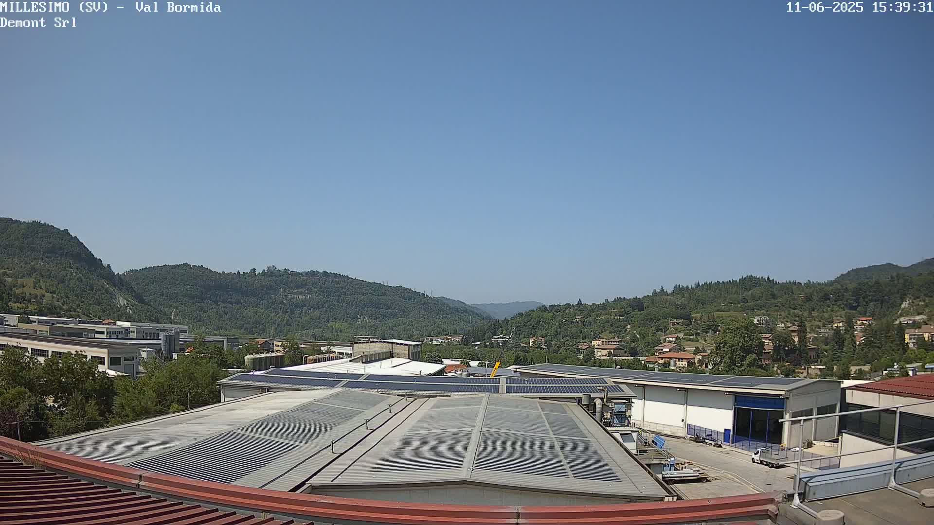 Under a clear blue sky, a view overlooking industrial buildings with solar panels on their roofs stretches towards a valley filled with green, tree-covered hills.
