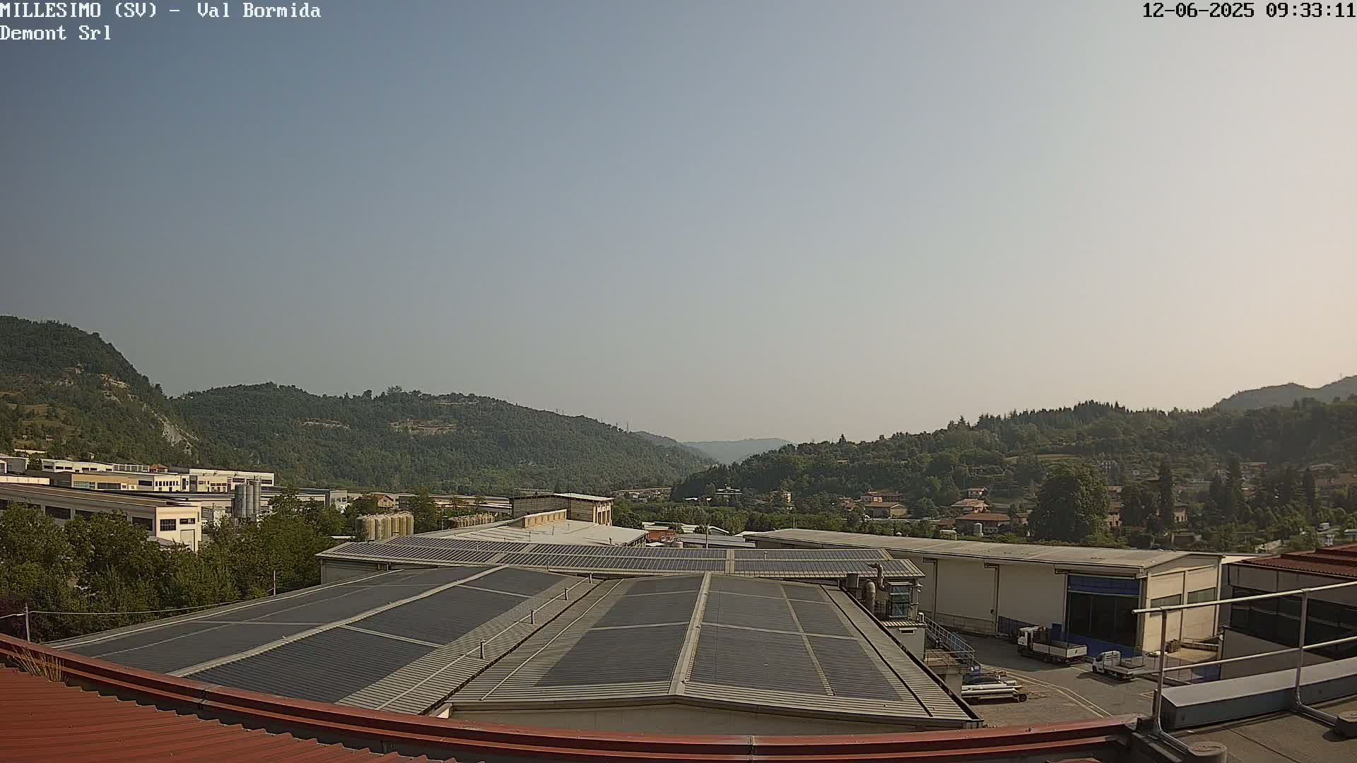 A view from a rooftop shows a factory complex with solar panels, surrounded by green hills under a clear sky.