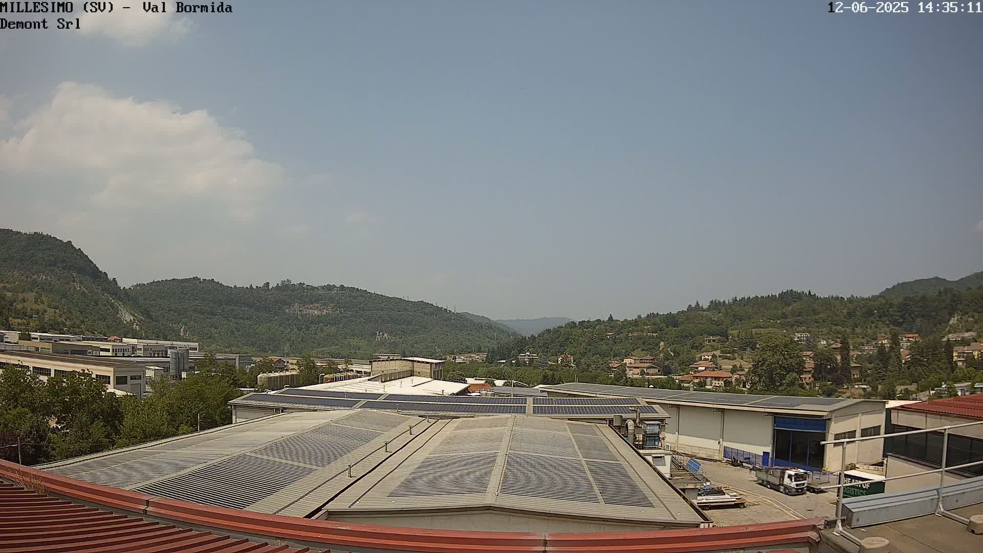 A mostly sunny view of industrial buildings with solar panels on their roofs, situated in a valley surrounded by green hills.