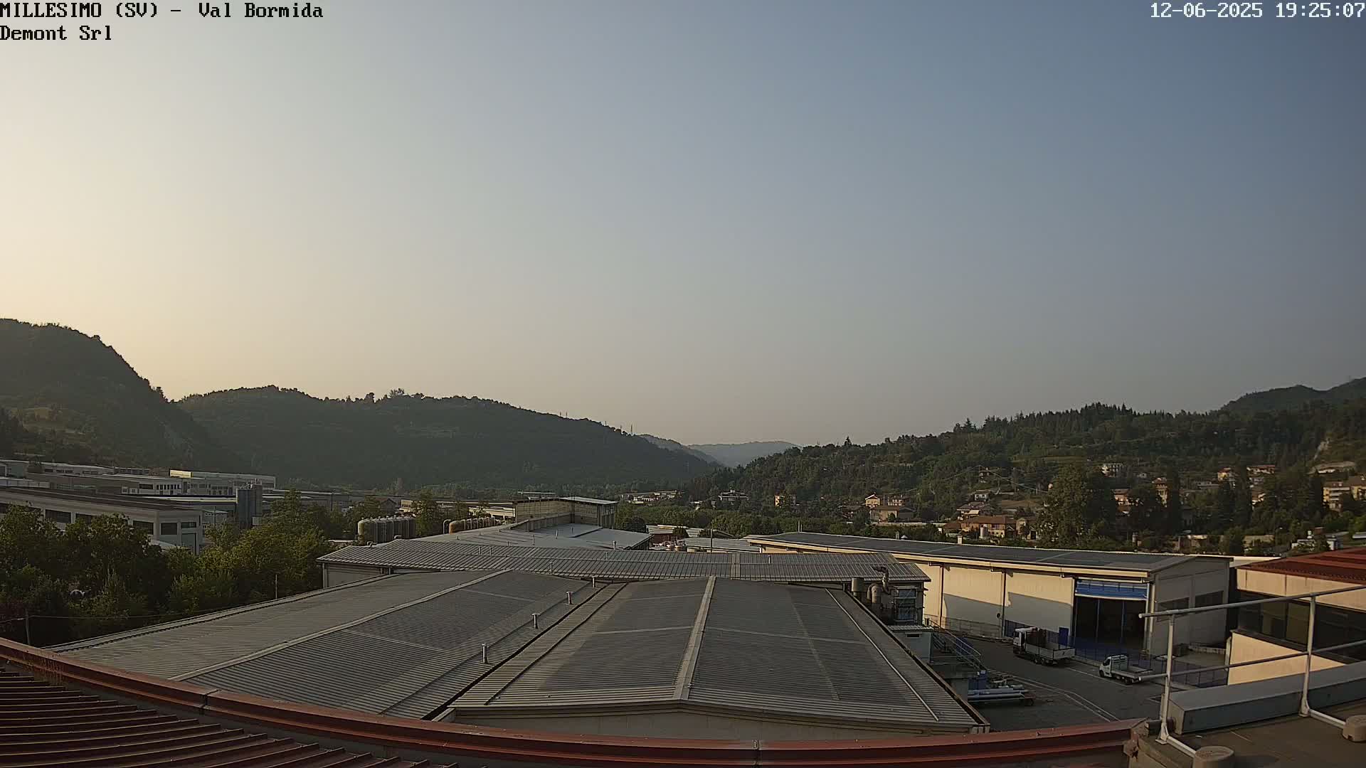 A view from a rooftop shows industrial buildings and a valley with hills and a town under a clear, light-blue sky.