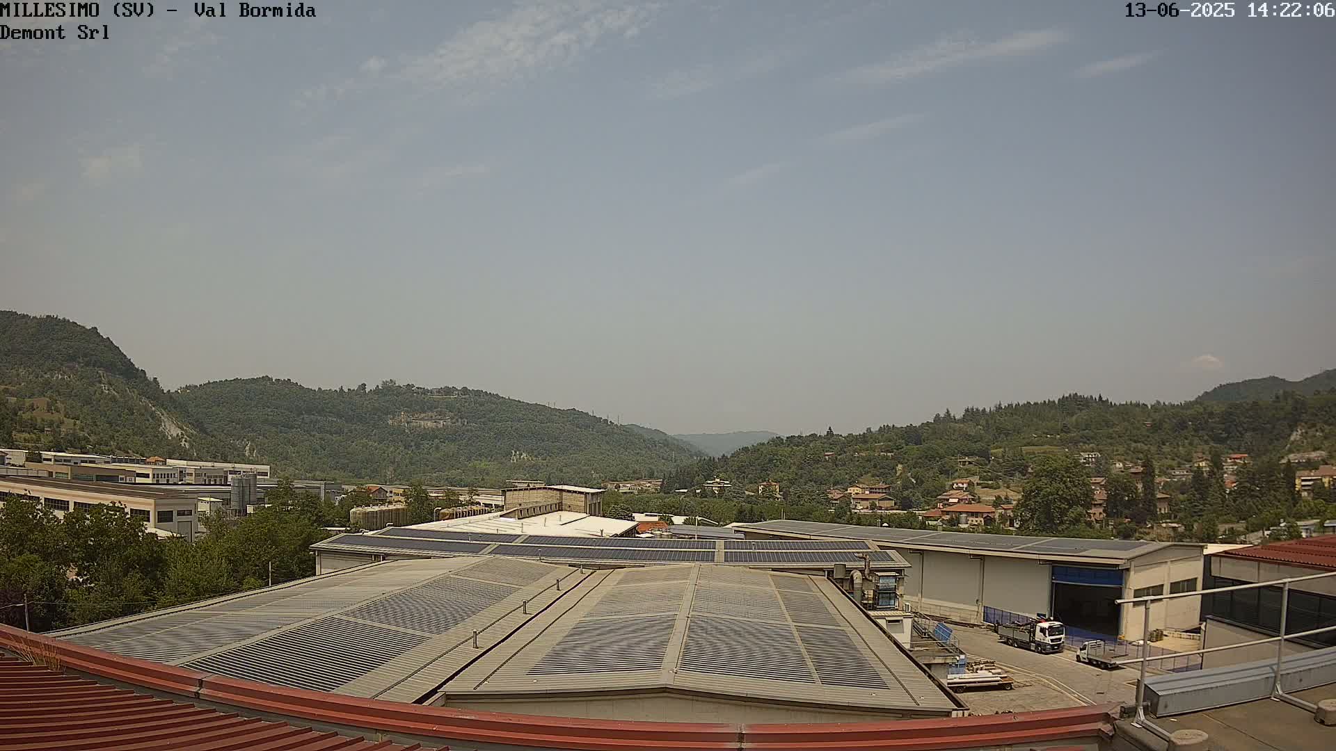 A mostly sunny day reveals a valley of green hills and a factory complex with solar panels on its roof.