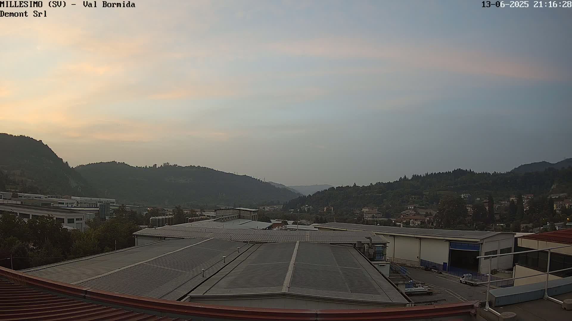 A hazy twilight sky over a valley of factories and residential buildings nestled among rolling green hills.