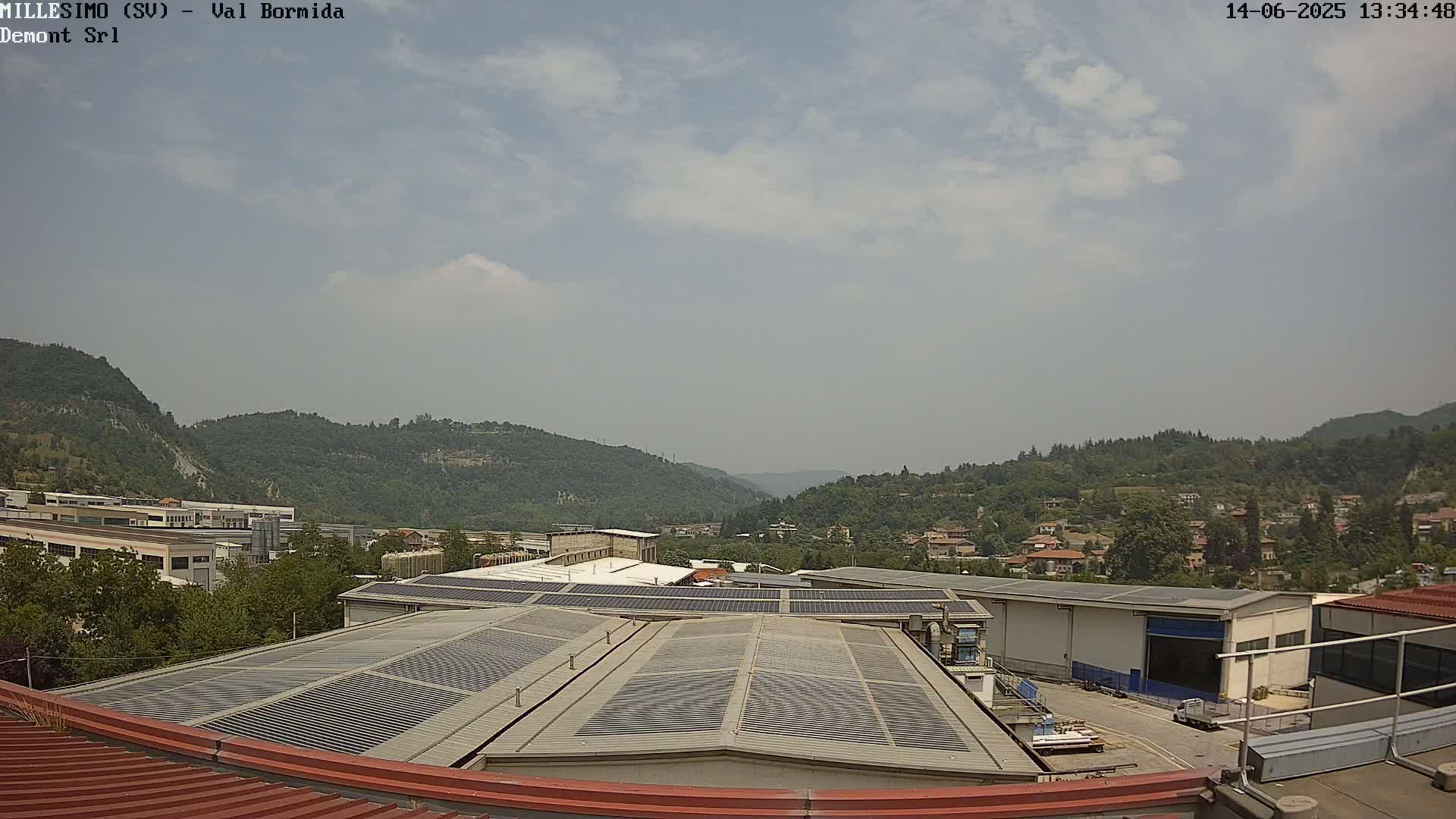 A view from a rooftop shows industrial buildings with solar panels, nestled in a valley surrounded by green hills under a partly cloudy sky.