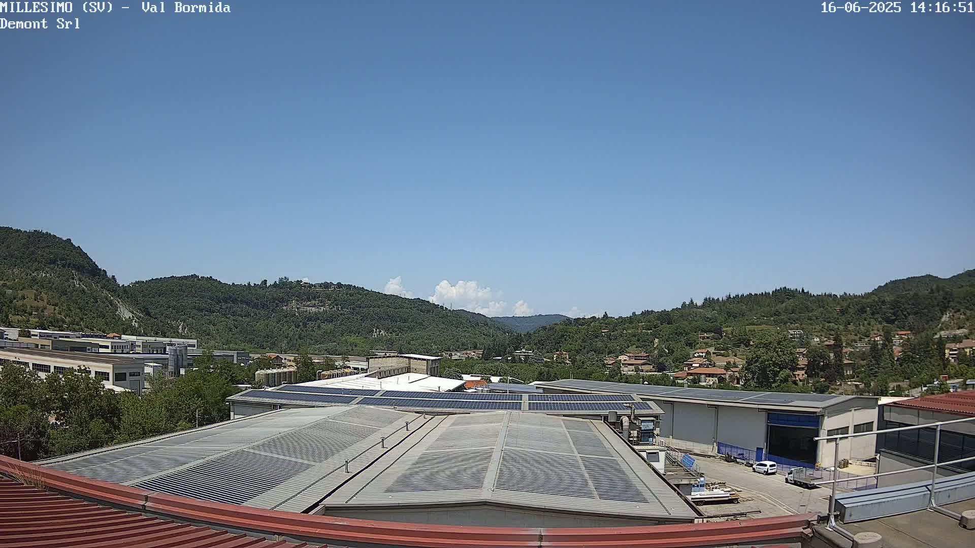 A clear blue sky overlooks a valley of green hills and a town, with industrial buildings featuring solar panels on their roofs in the foreground.