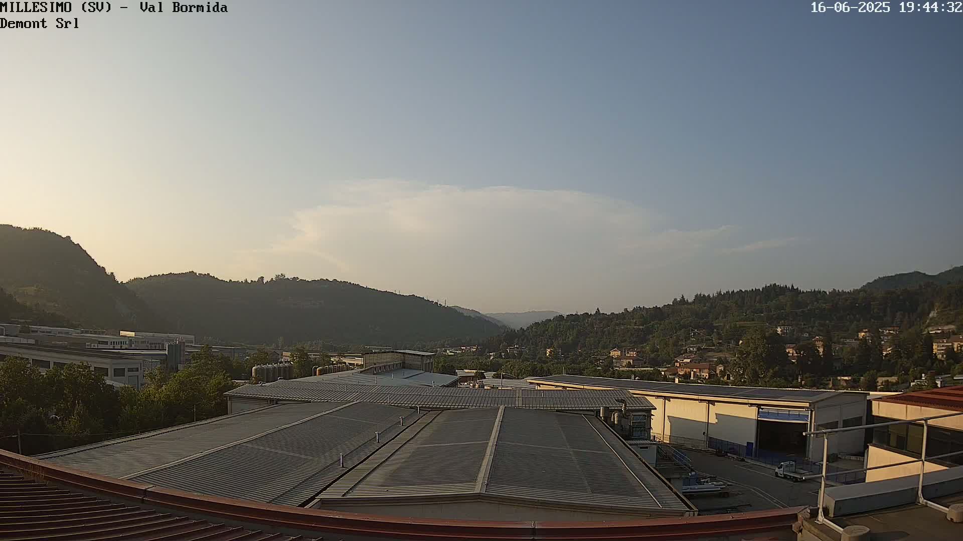 A view from a rooftop shows a mostly clear sky, industrial buildings, and tree-covered hills in the distance.