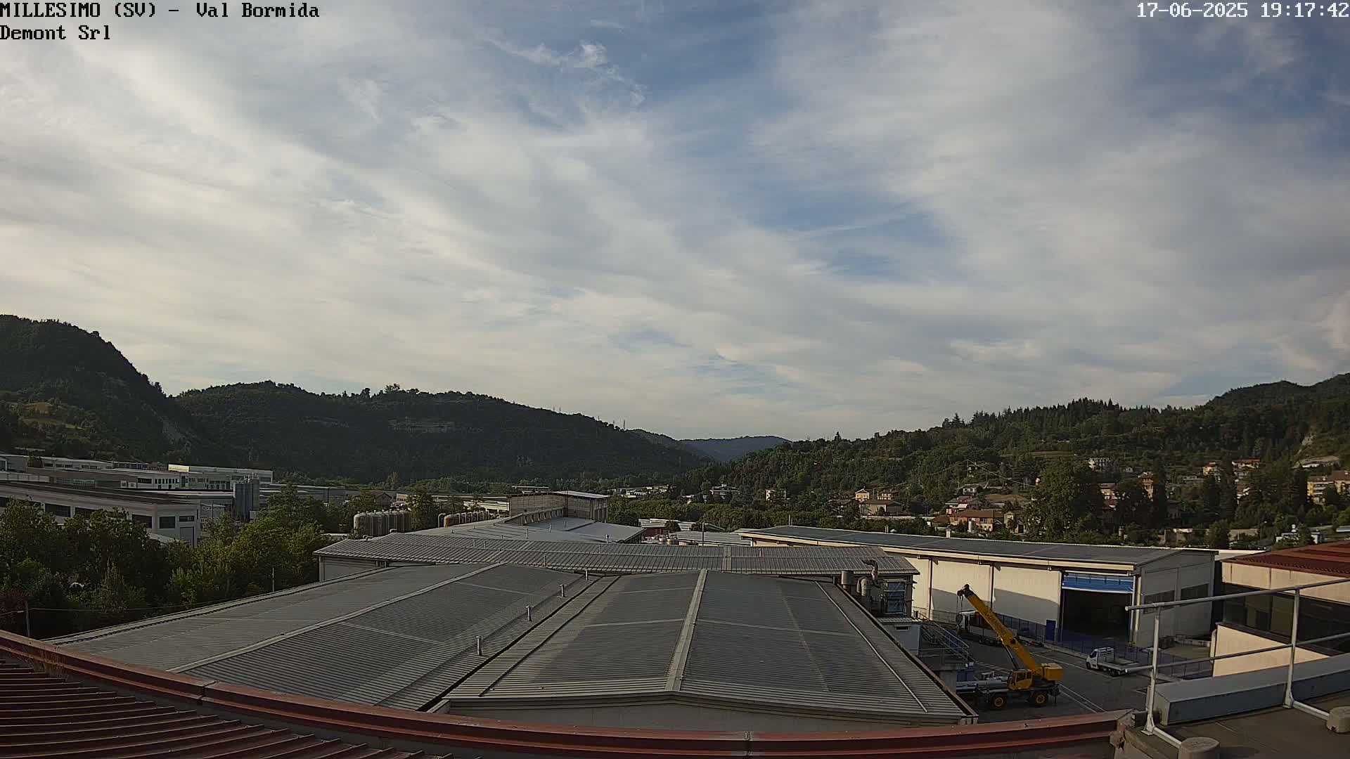 A partly cloudy sky overlooks a factory complex nestled in a valley between green hills.