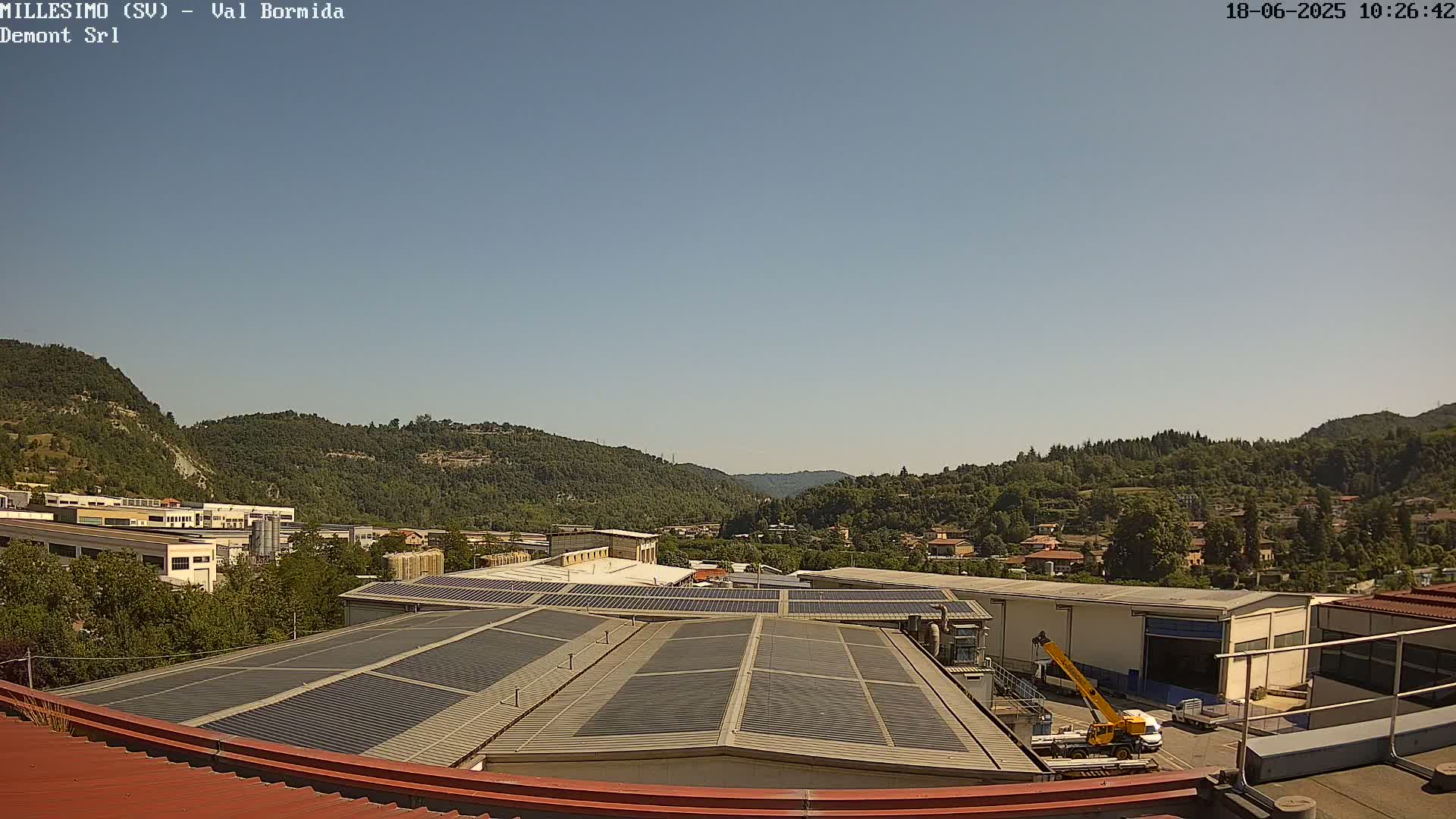 A clear, sunny day reveals a view of industrial buildings with solar panels on their roofs, nestled among green hills and a small town in the distance.