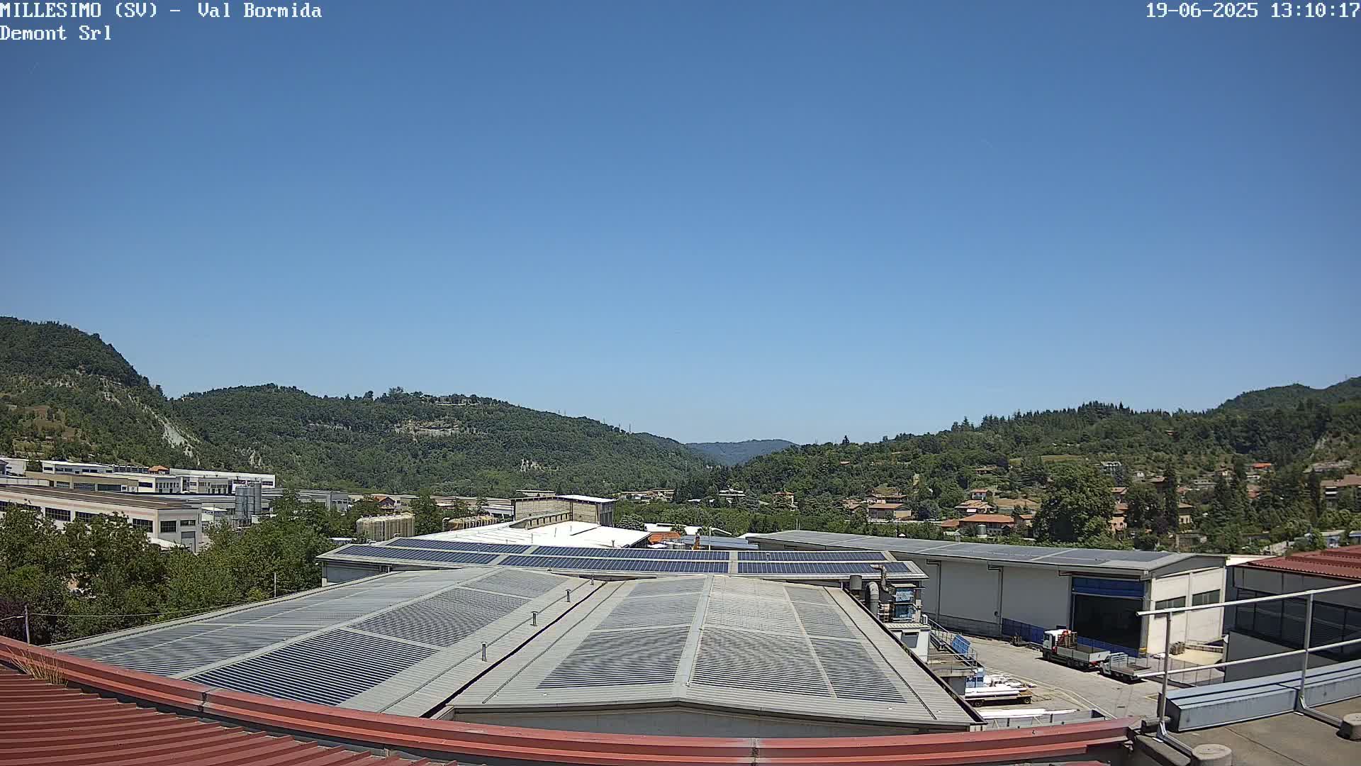 A clear blue sky overlooks a valley of green hills and industrial buildings with solar panels on their roofs.